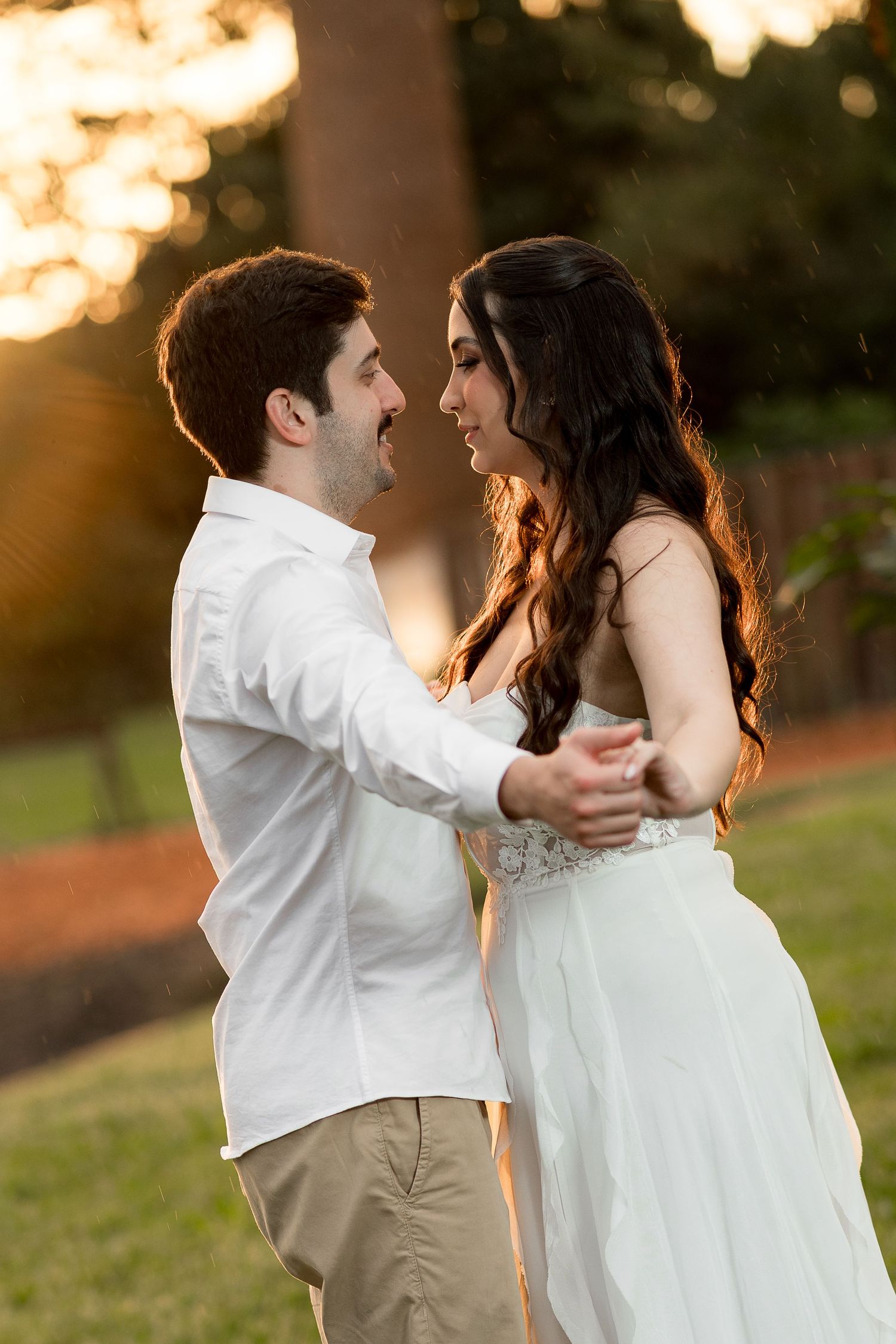A couple in white attire dancing and smiling at each other outdoors during golden hour sunset.