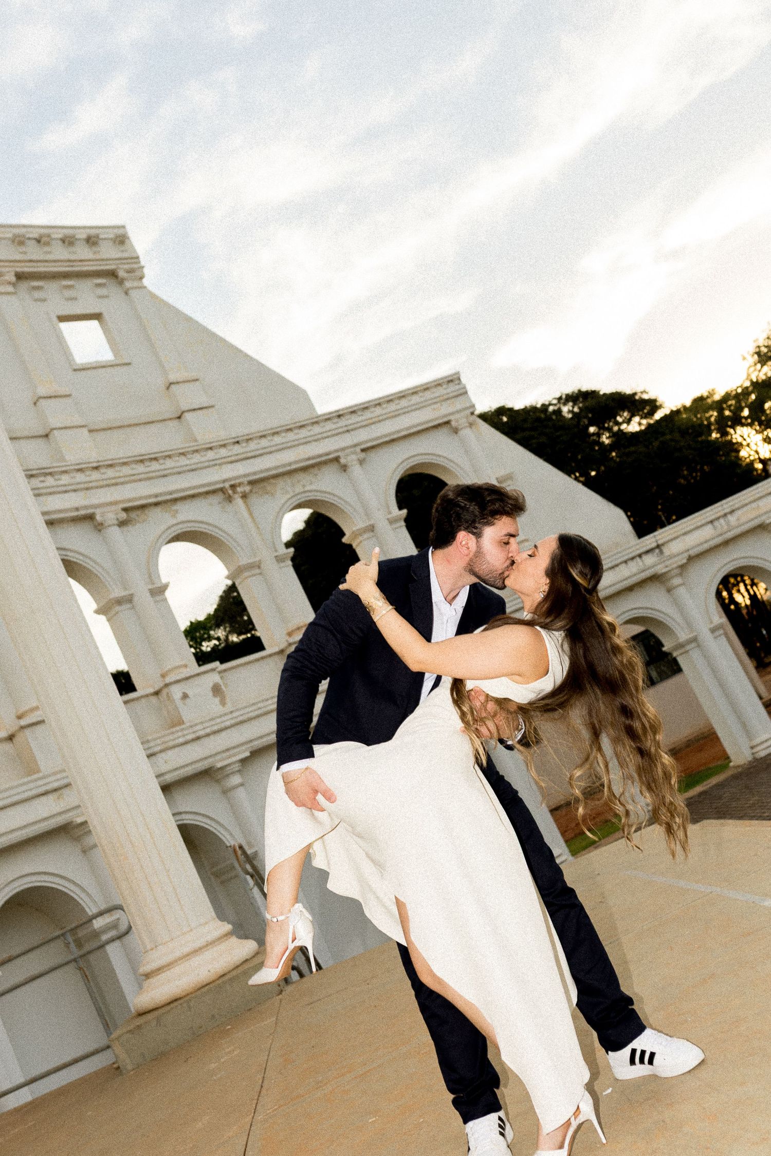 Couple sharing a romantic kiss dip in front of a white classical arch monument at golden hour.