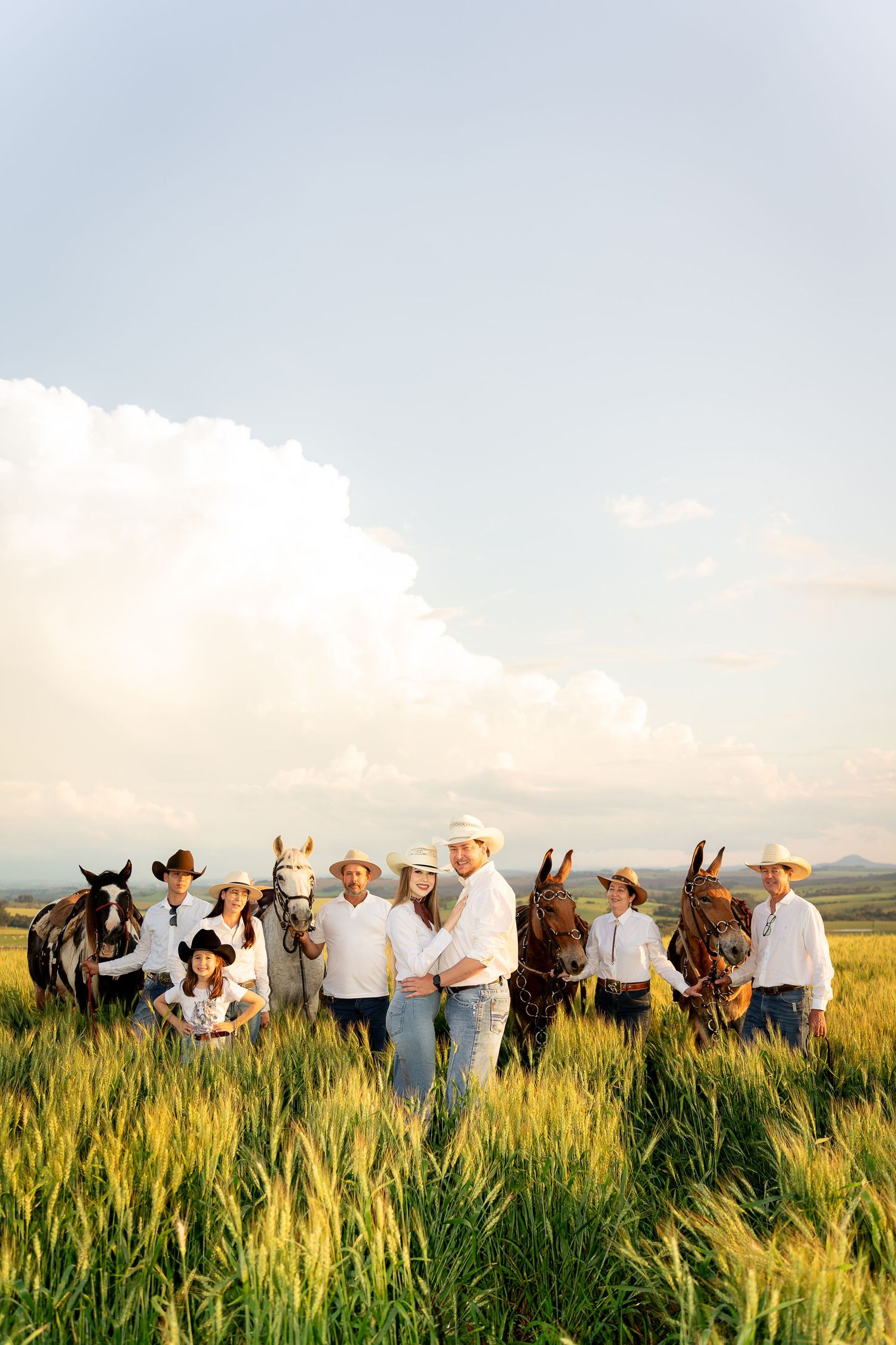 A family in white shirts and cowboy hats poses with horses in a sunlit green field under a cloudy sky.