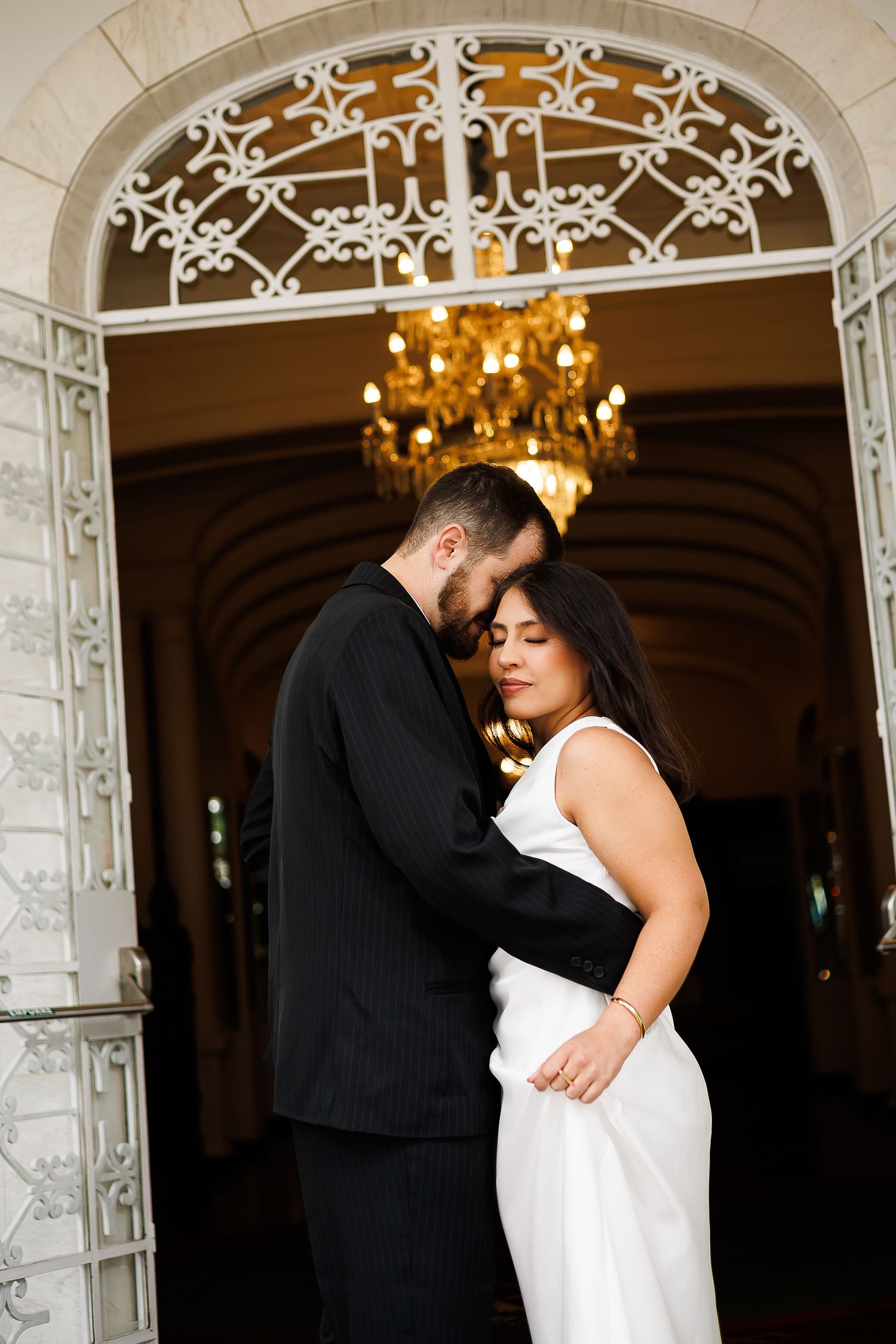 Elegant couple embracing in front of ornate white iron doors with glowing chandelier, bride in white gown and groom in black suit.