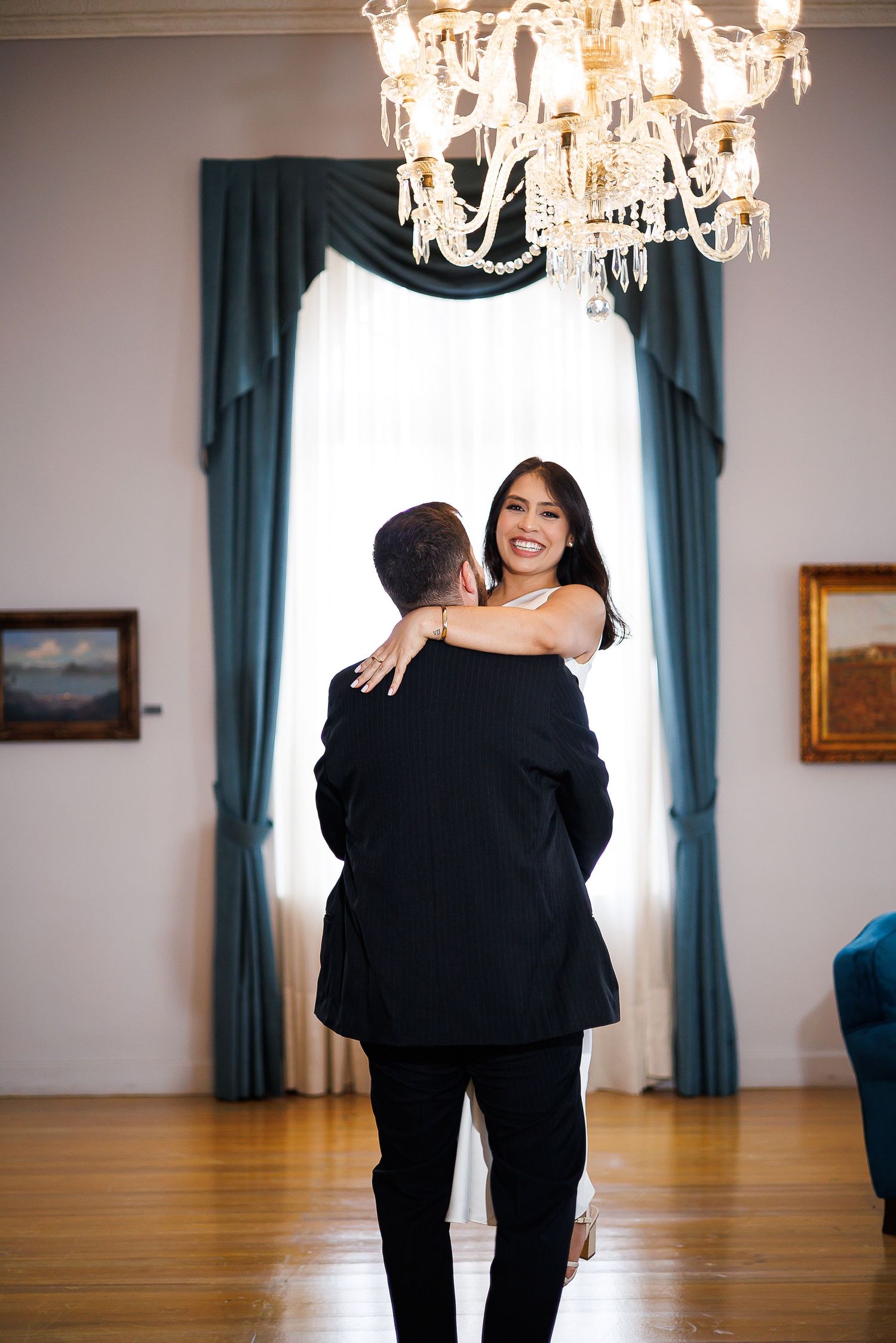 Couple dancing elegantly in a grand room with crystal chandelier, teal drapes, and hardwood floors.