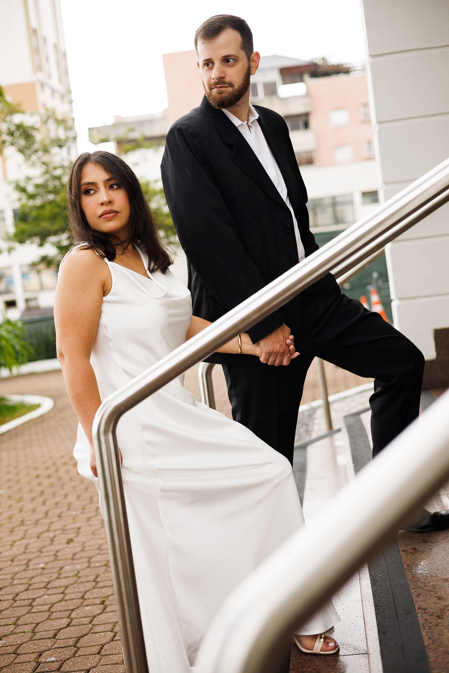 Bride in white gown and groom in black suit holding hands on outdoor stairs in urban setting.
