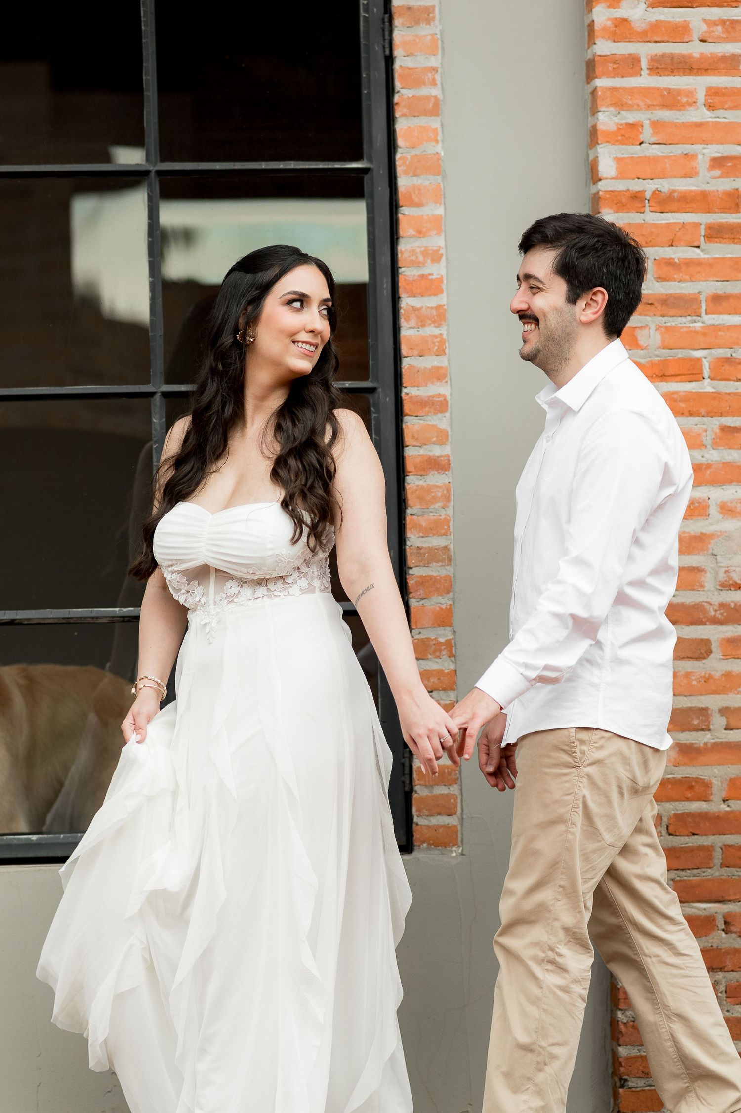 A smiling bride in a white gown holds hands with a groom in a white shirt near a brick wall.