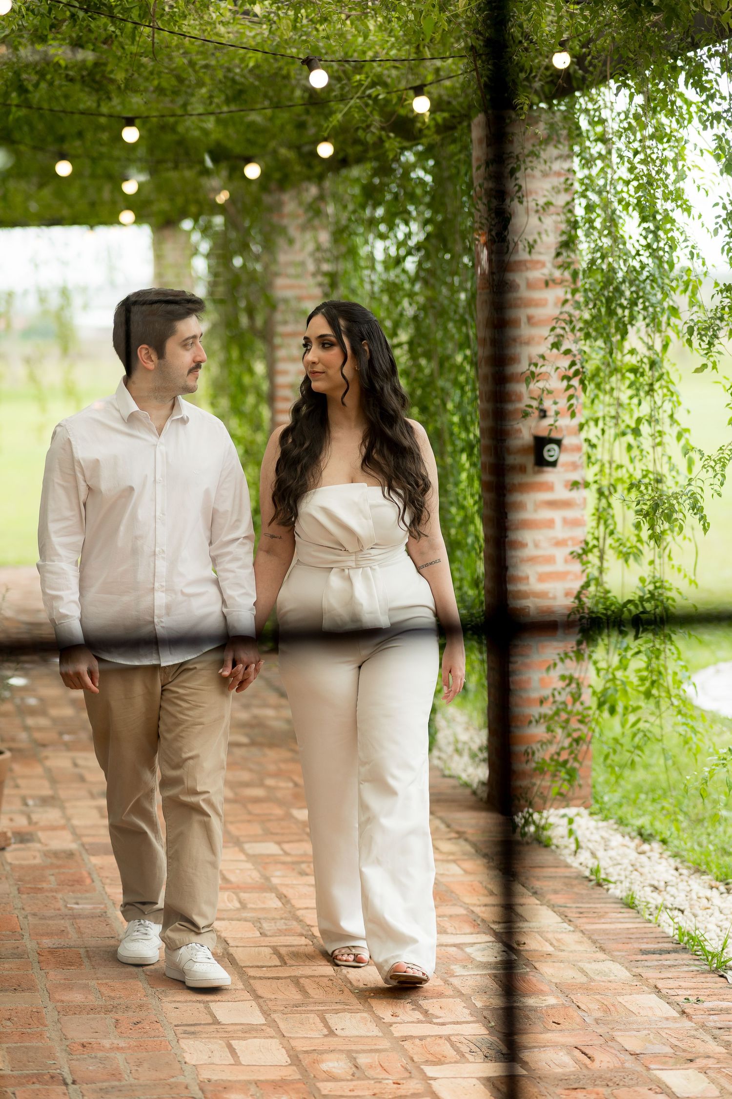 Couple walking hand in hand under string lights on a brick path surrounded by lush greenery.