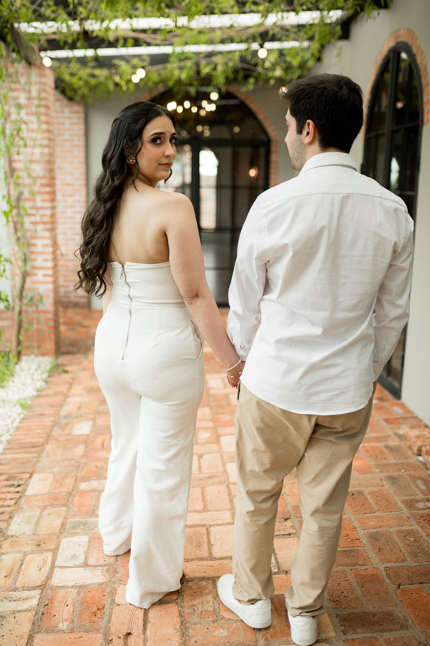 Couple holding hands walking through brick courtyard, woman in white strapless jumpsuit glancing back, man in white shirt.