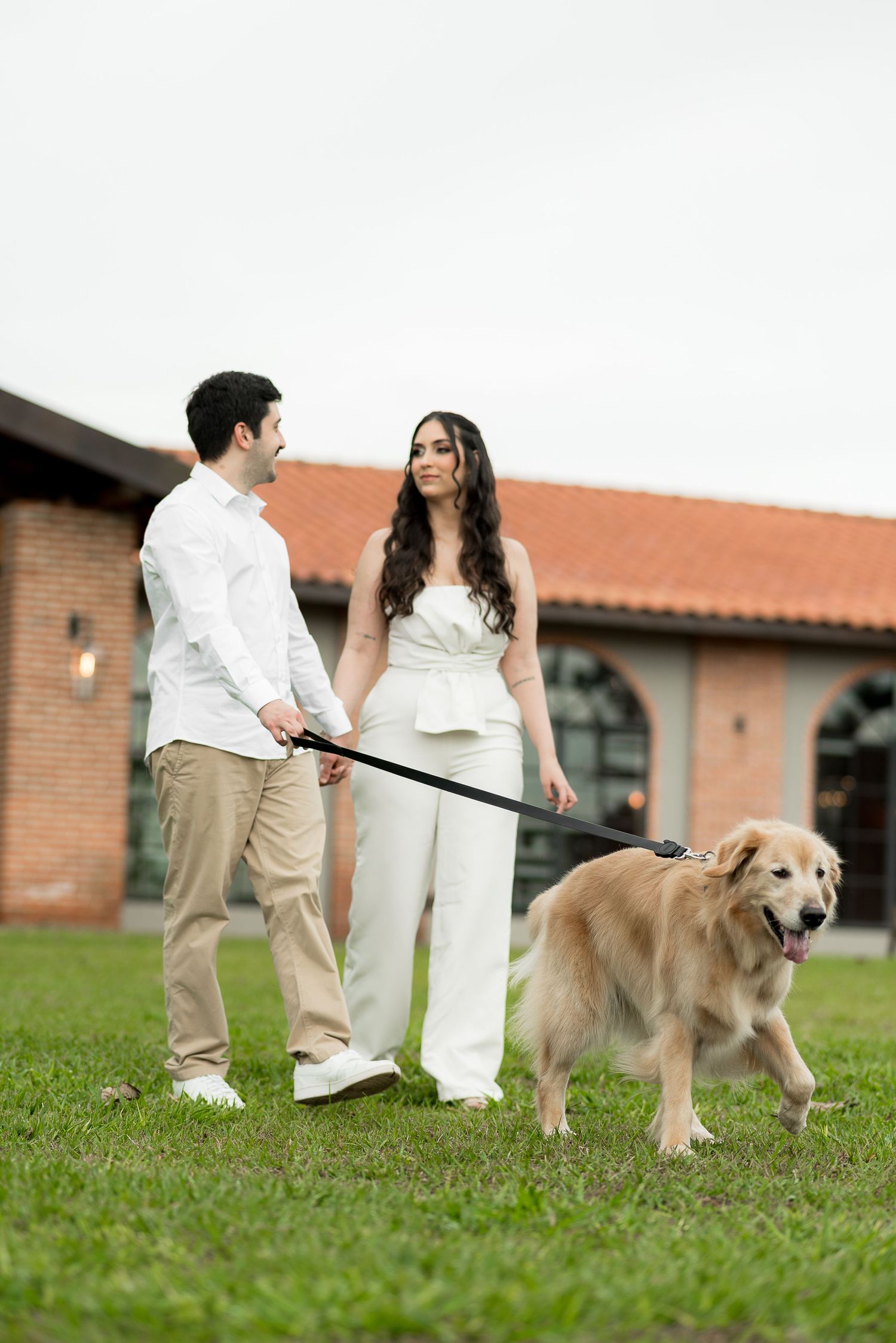 Couple in white and khaki walking a golden retriever on a leash outside a brick building on green grass.