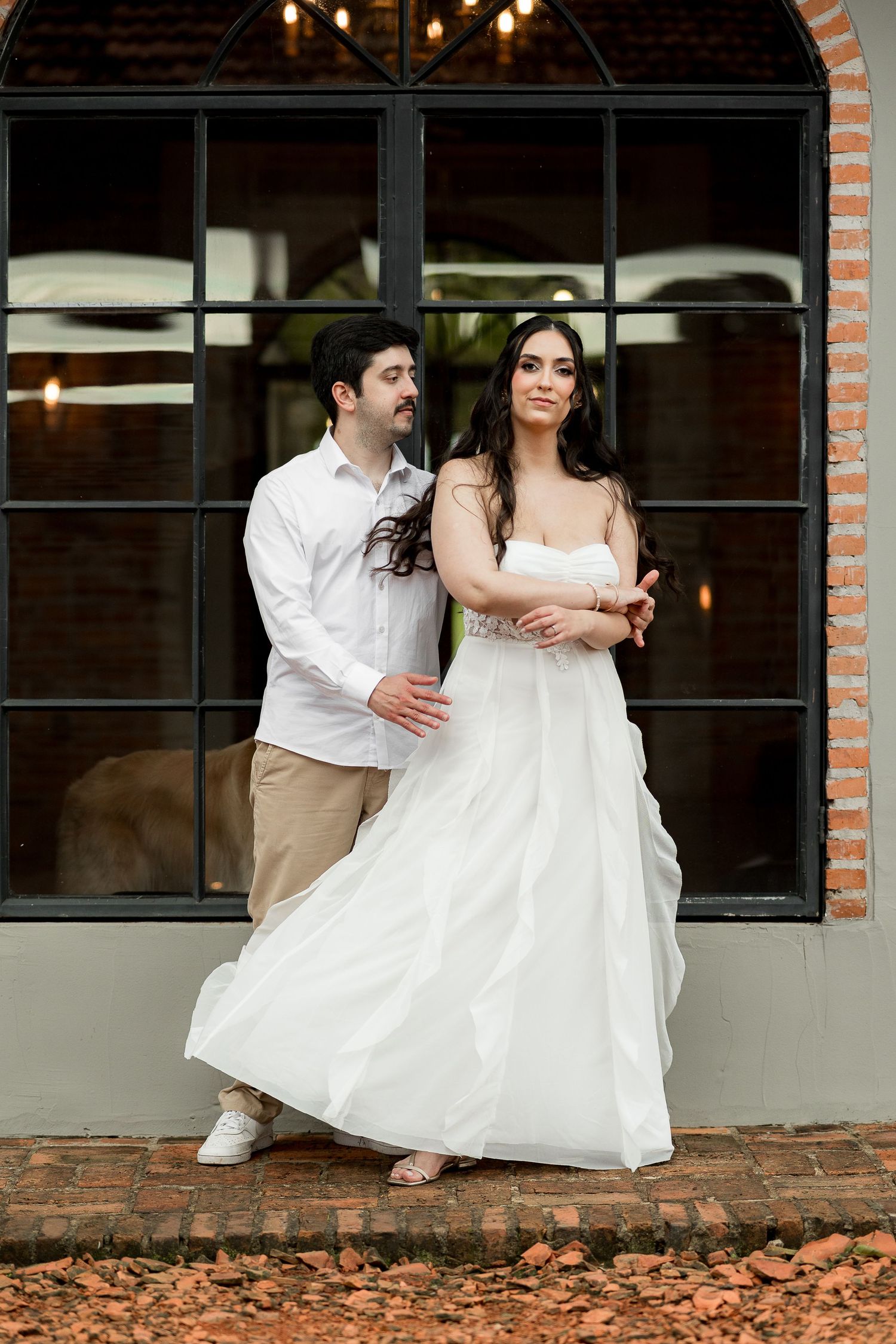 Couple in wedding attire posing outside a rustic venue, bride in white gown and groom in white shirt and khakis.