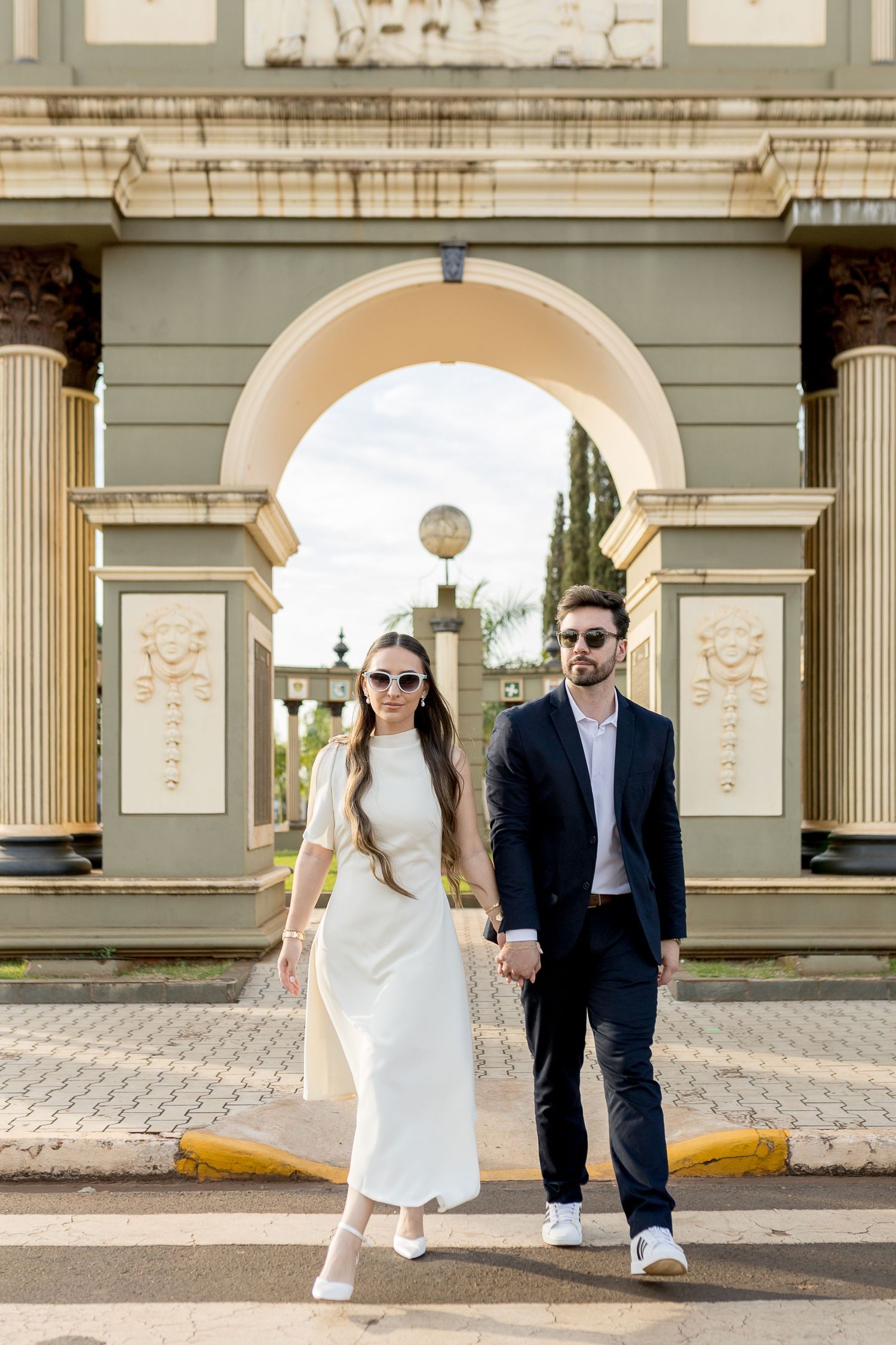 Elegant couple in sunglasses holding hands walking under a grand arch, woman in white dress, man in dark suit.