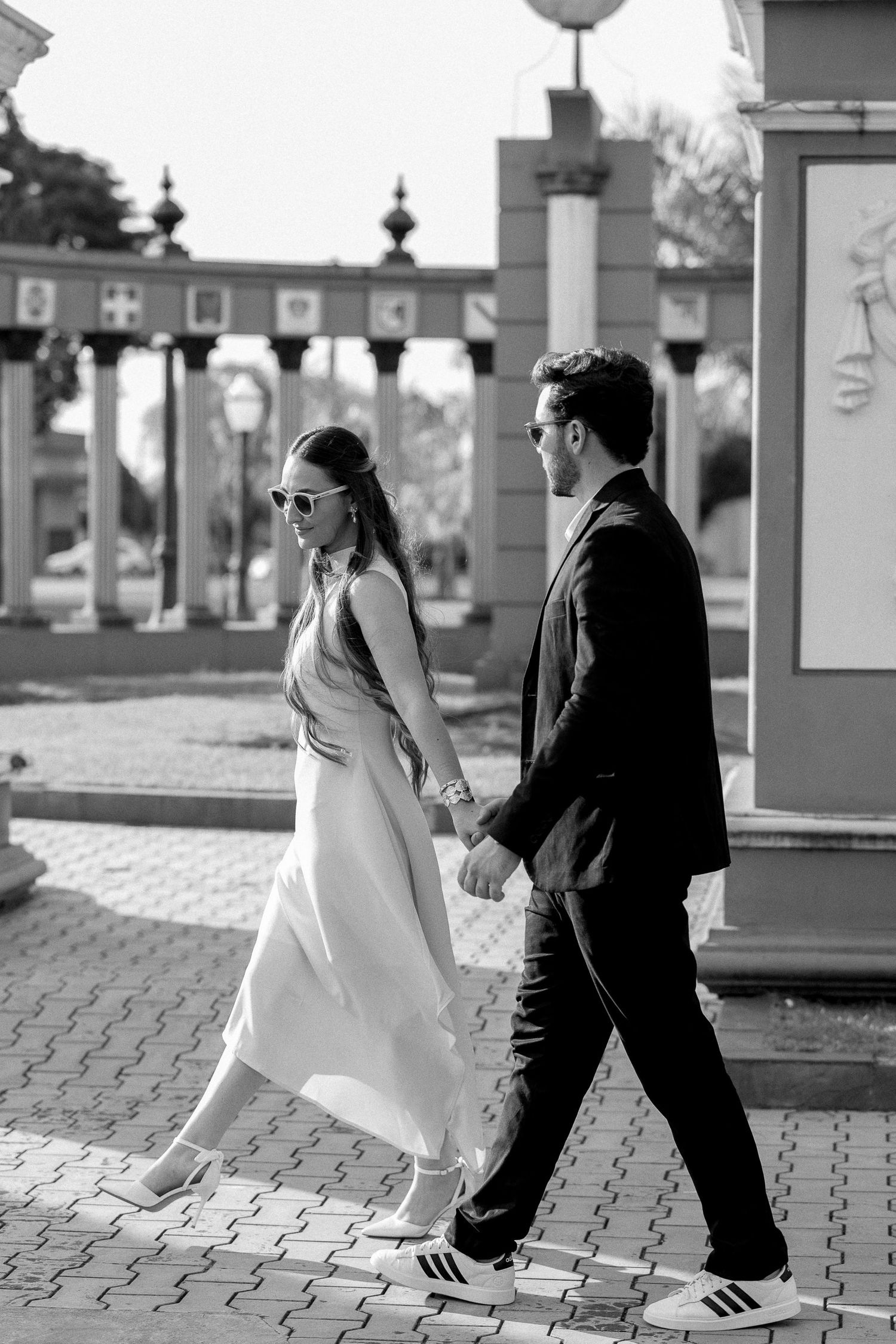 Black and white photo of stylish couple holding hands walking past ornate columns outdoors.