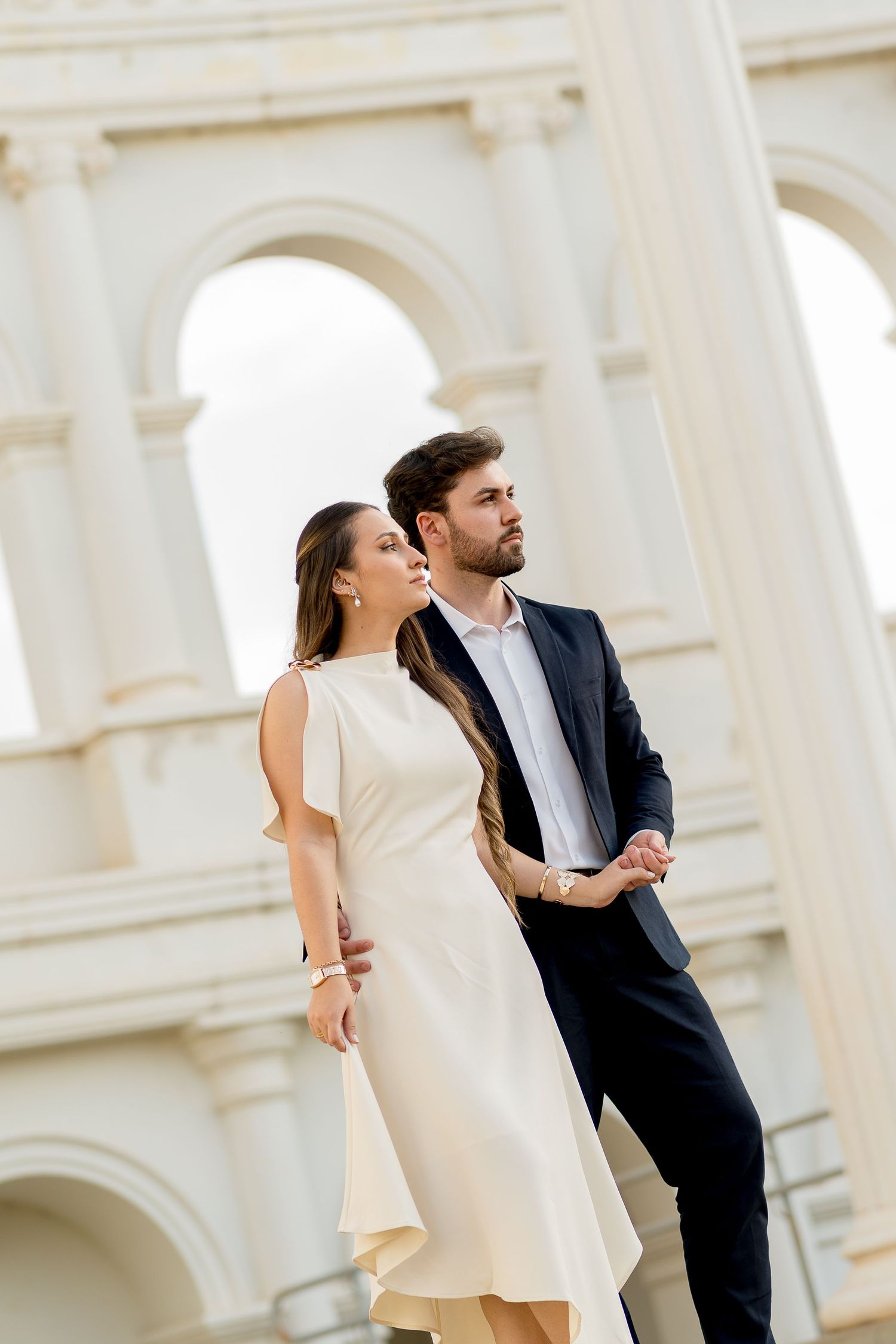 Elegant couple posing in front of classical white arched building, woman in cream dress, man in navy suit.
