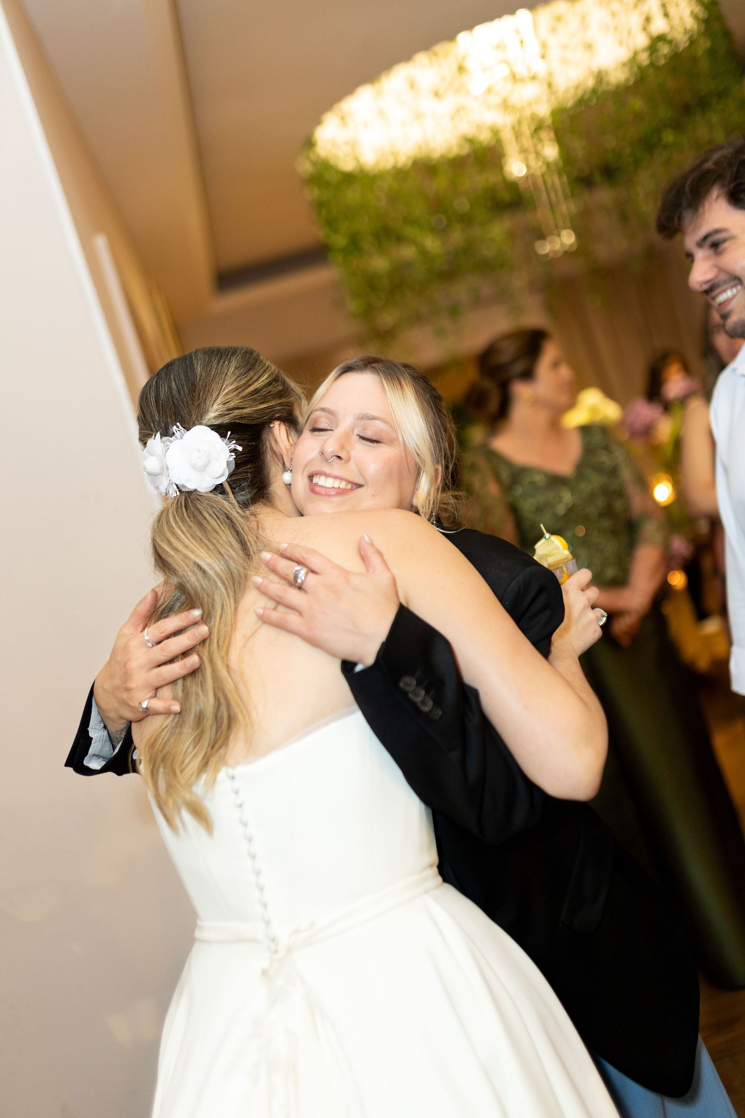Bride in white gown with floral hair accessory shares a joyful hug with a smiling blonde guest at a wedding reception.