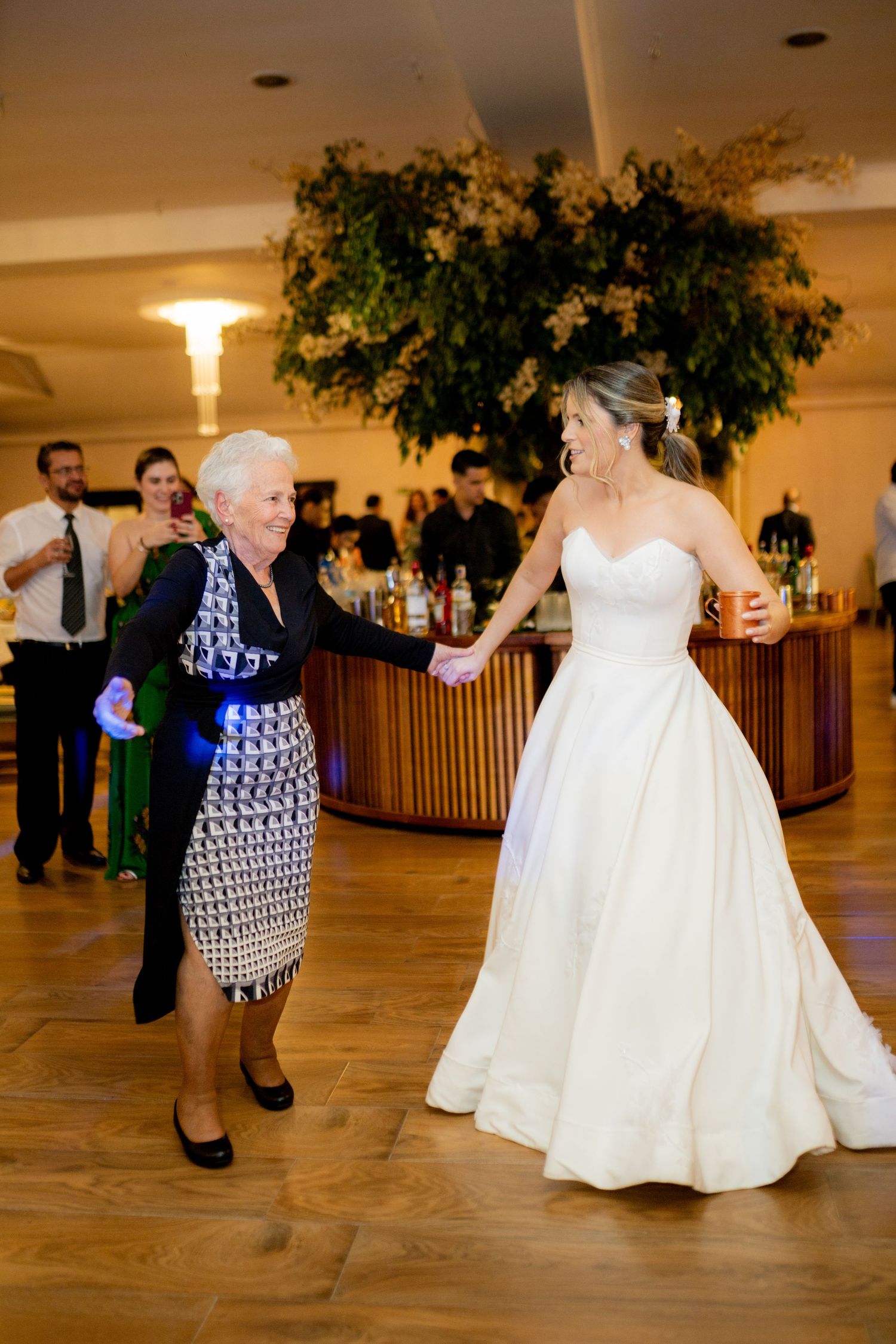 Bride in white gown dancing joyfully with elderly woman in patterned dress at elegant wedding reception.