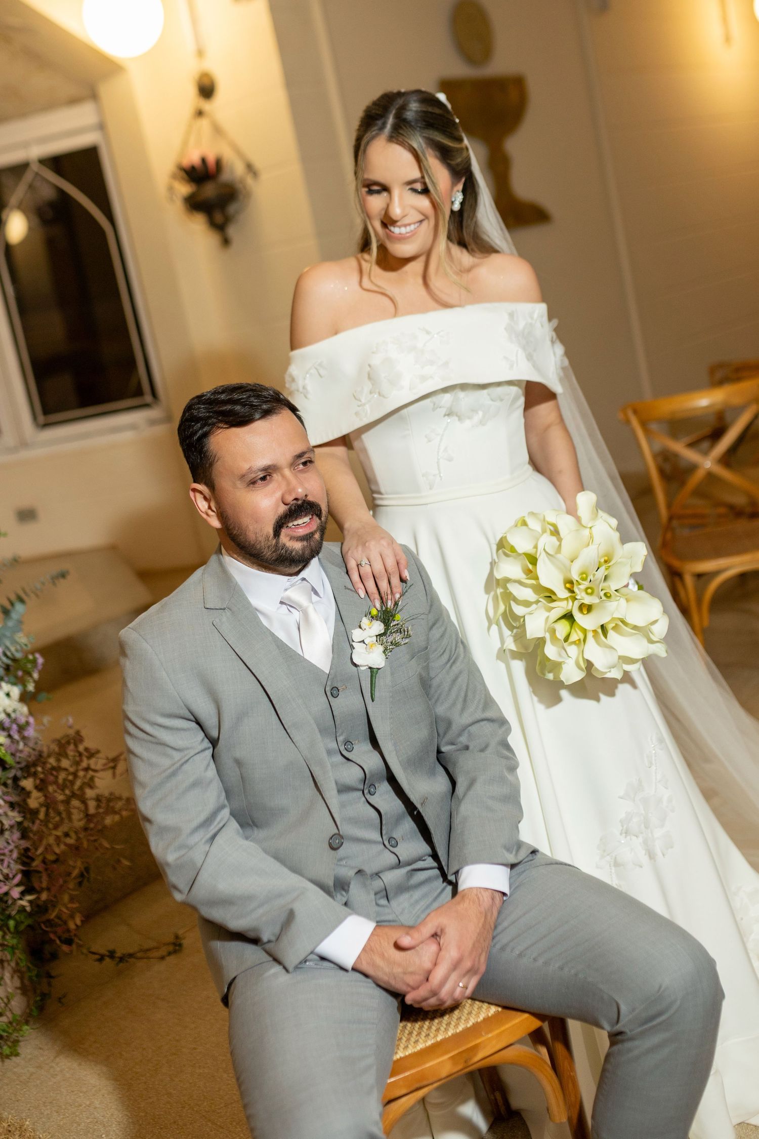 Bride in white off-shoulder gown holding calla lily bouquet stands behind groom in gray suit seated at wedding ceremony.