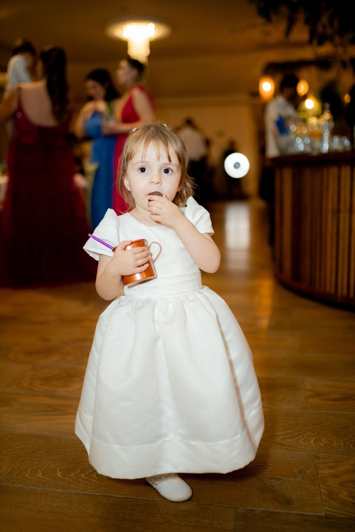 Young flower girl in white dress holding a mug at a wedding reception venue.