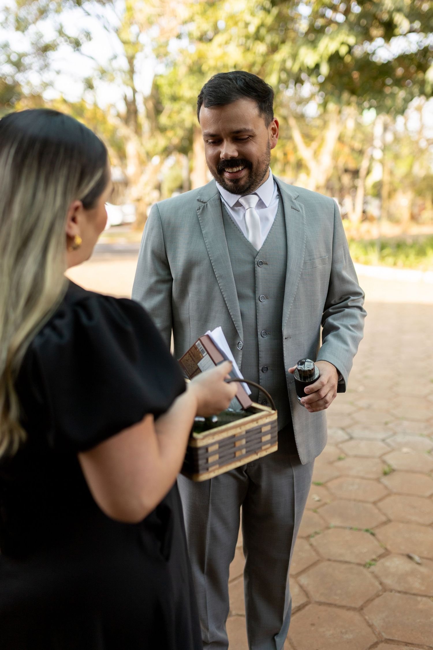 A smiling man in a gray suit holds a wine glass while a woman in black dress carries a wooden box outdoors.