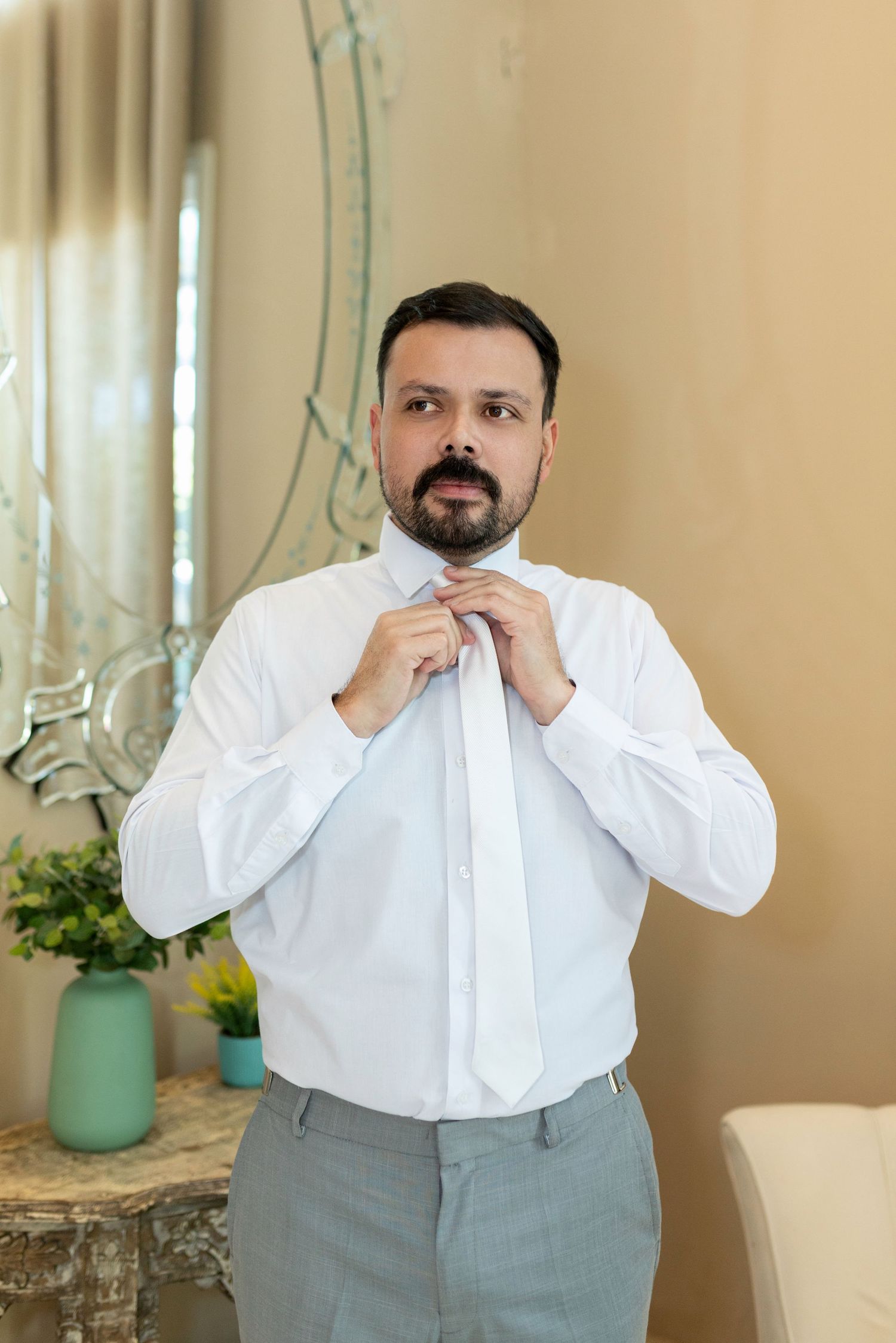Bearded man in white dress shirt adjusting white tie while getting dressed in elegant room.