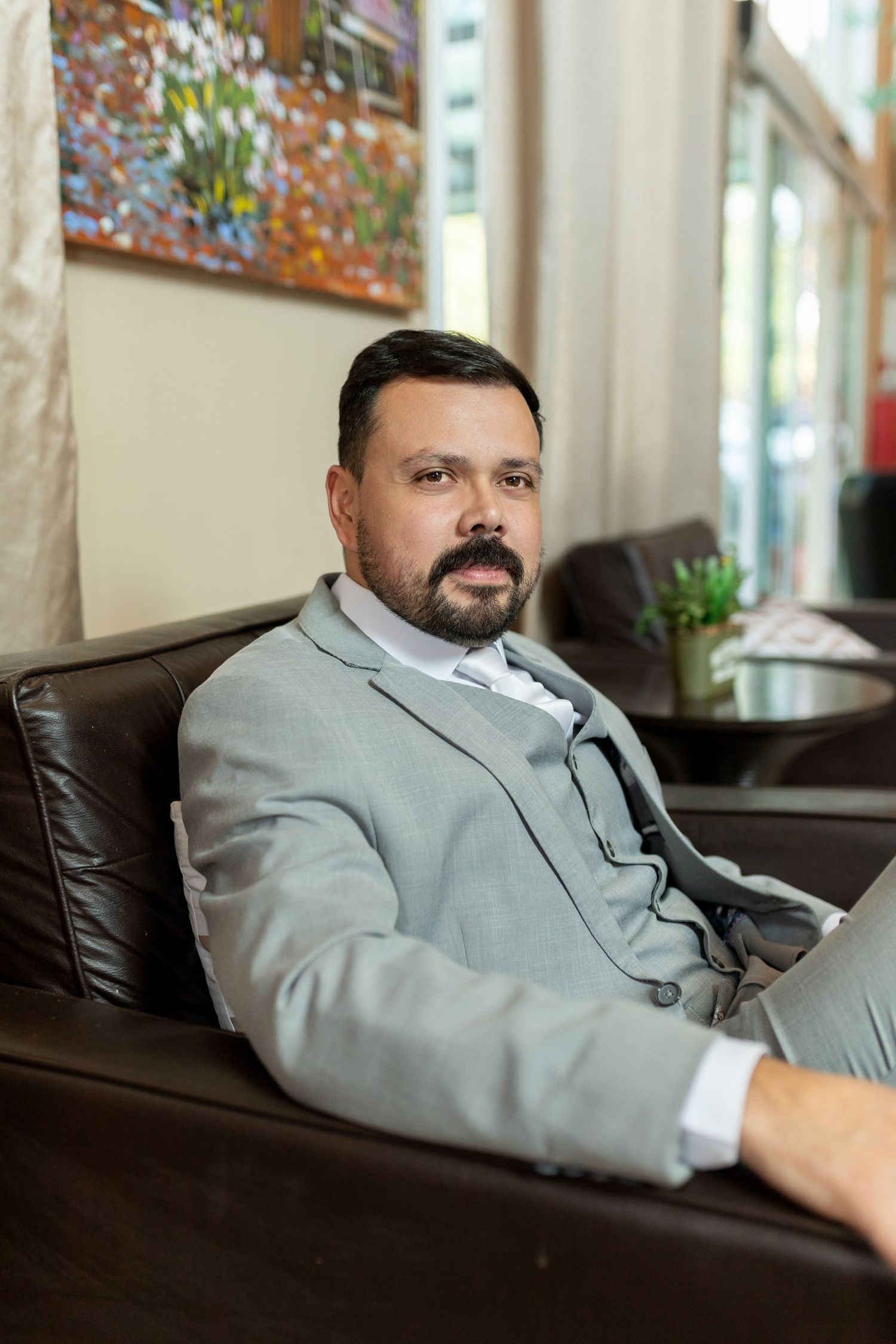 Professional man in gray suit sitting relaxed on leather chair in modern office lobby with artwork on wall.