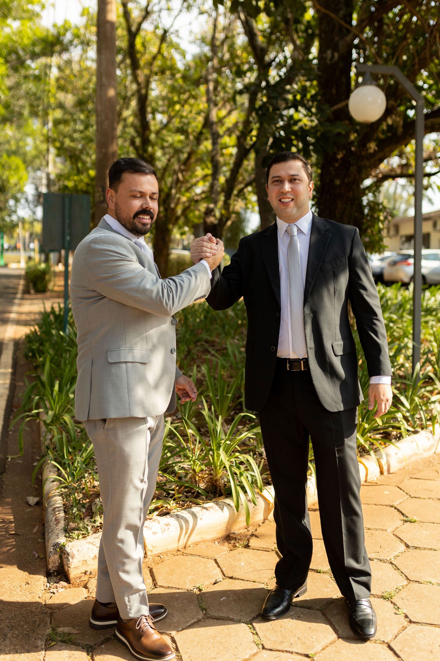 Two businessmen in suits shake hands outdoors in a park setting, smiling warmly on a sunny day.