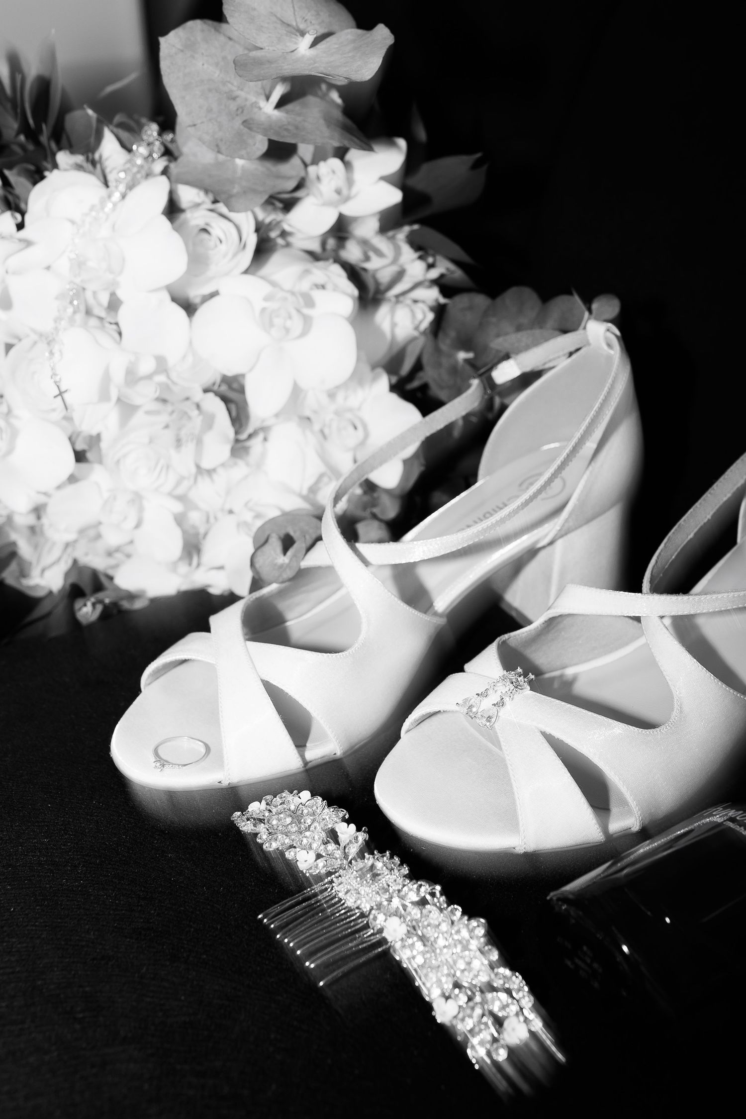Black and white photo of bridal heels, crystal hair comb, and white floral bouquet arranged elegantly for a wedding.