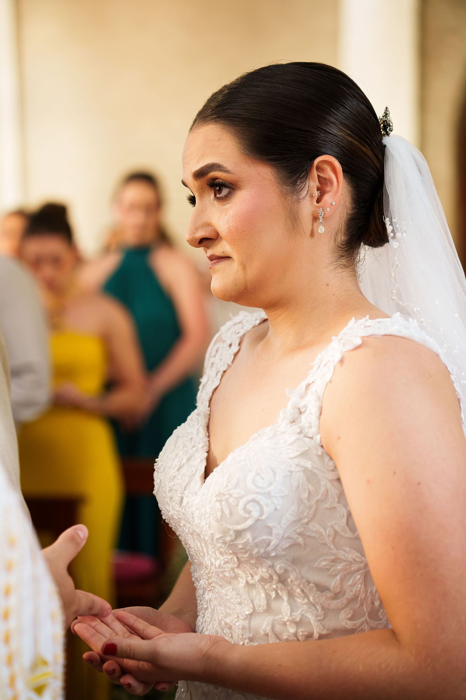 Bride in white lace gown and veil during wedding ceremony, with bridesmaids in colorful dresses in background.