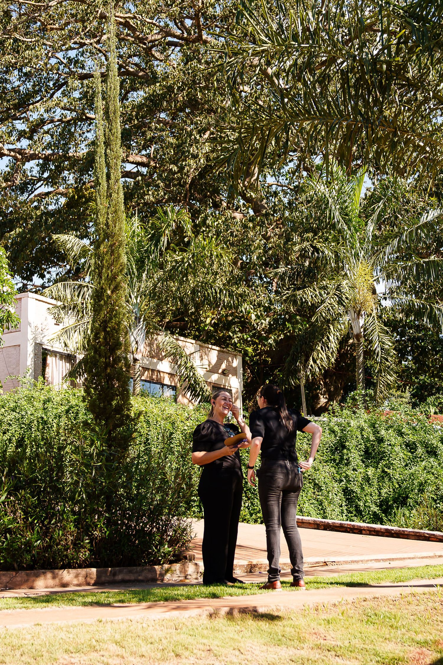 Two women in black outfits conversing in a lush green park surrounded by tall trees and manicured hedges.