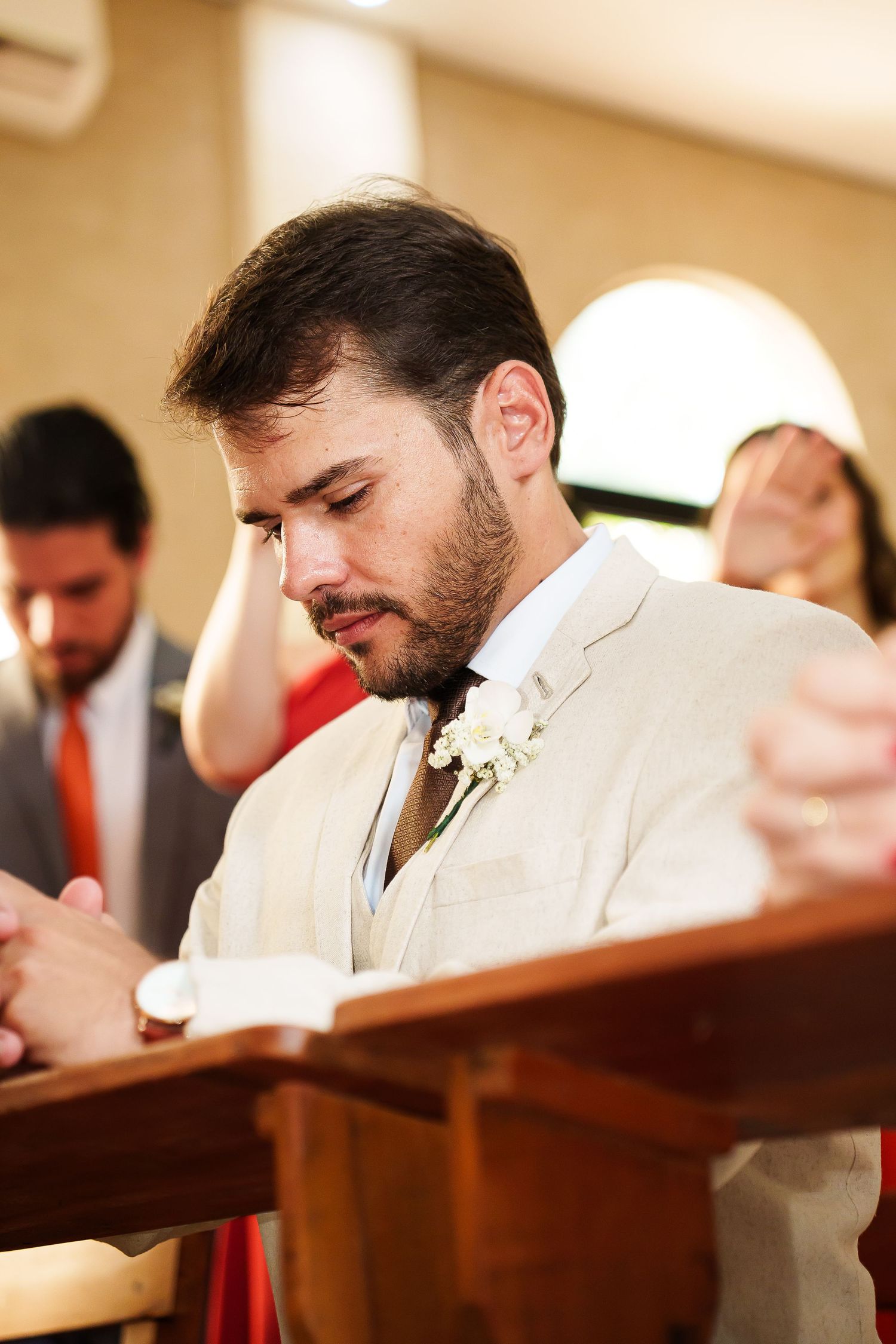 Groom in beige suit with boutonniere bows head in prayer at church pew during wedding ceremony.