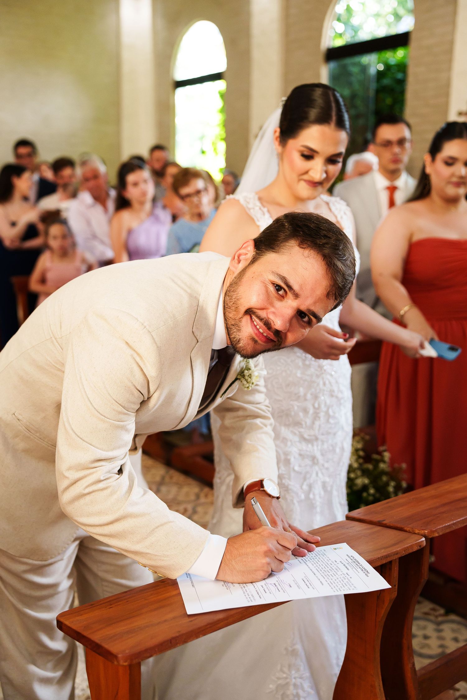 Smiling groom in beige suit signs wedding certificate at church altar with bride in white lace dress standing behind him.