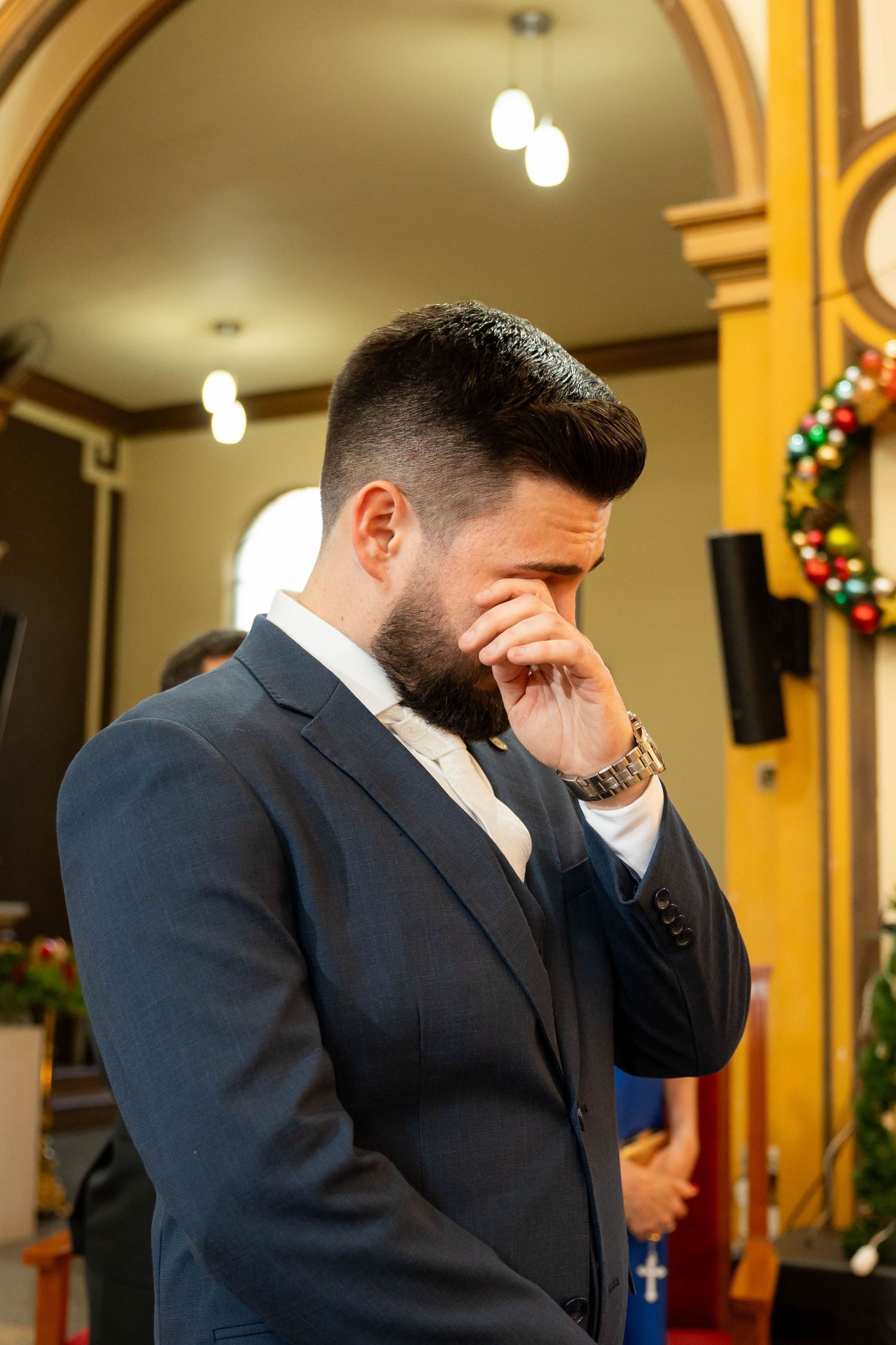 Emotional groom in navy suit wiping tears during wedding ceremony inside decorated church.