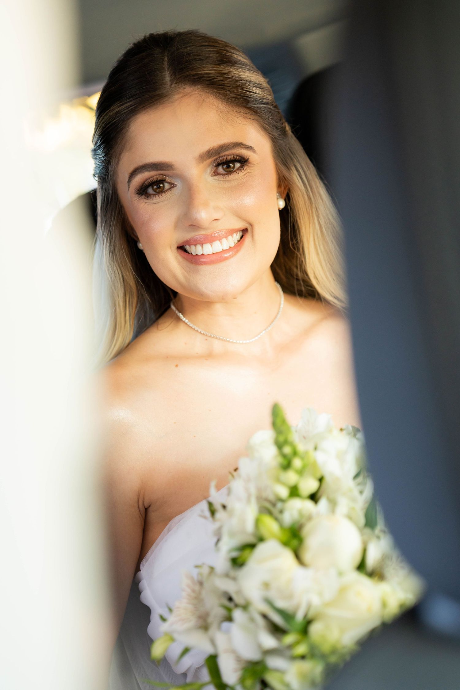 Smiling bride holding white flower bouquet, wearing strapless gown and pearl necklace on her wedding day.