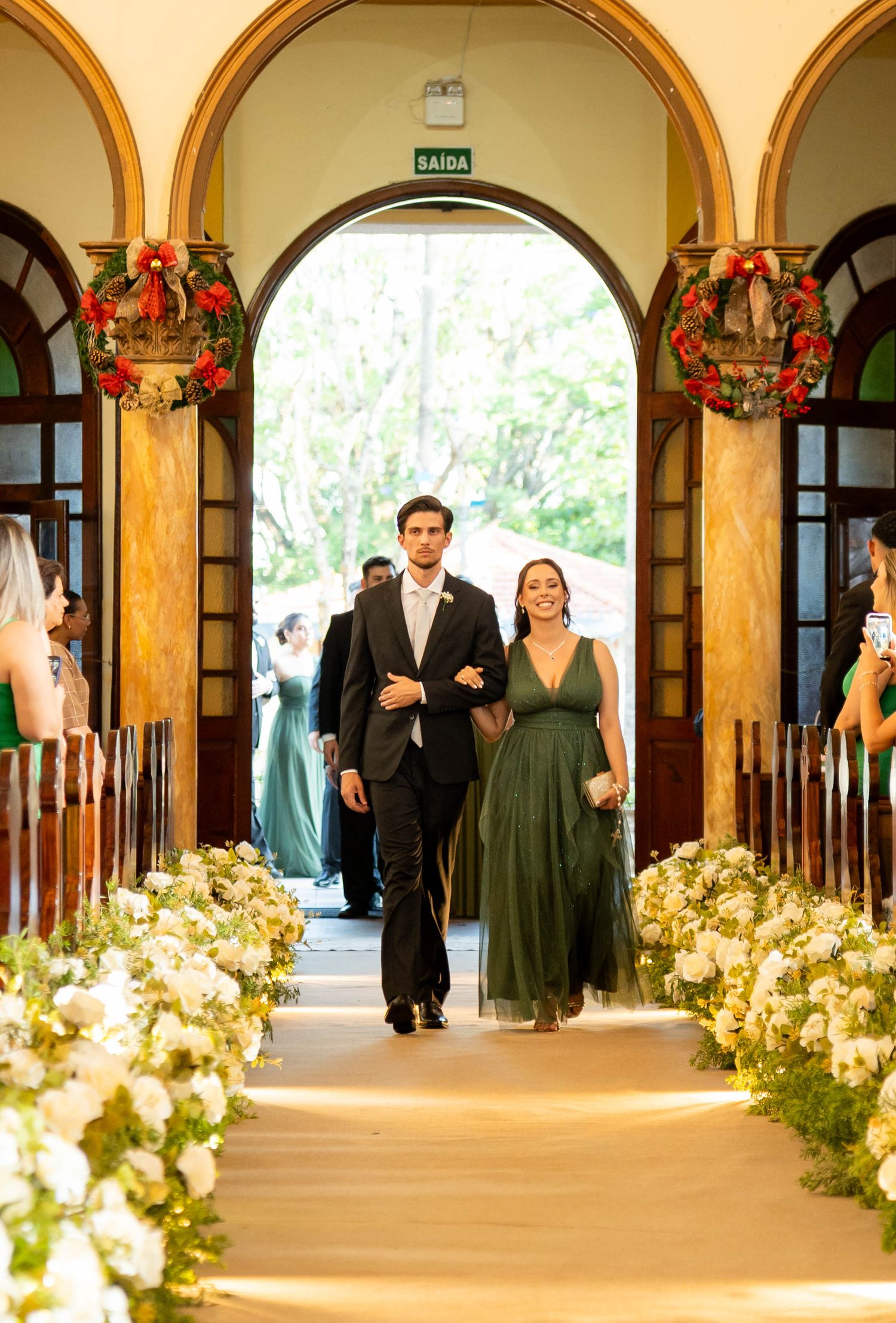Couple walks down floral-lined church aisle, woman in olive green gown, Christmas wreaths decorating entrance.