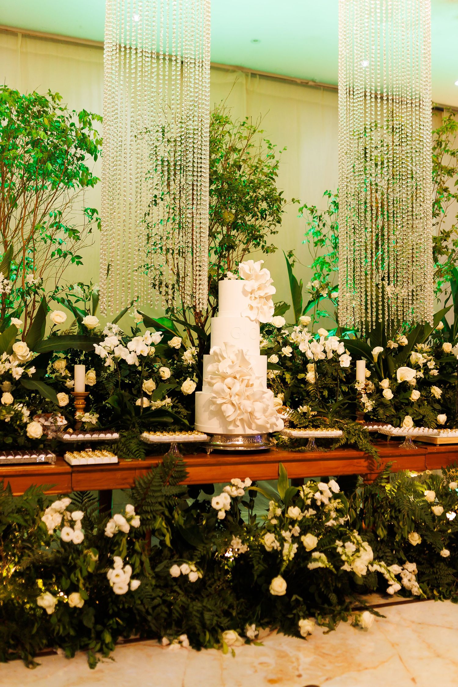Elegant white tiered wedding cake surrounded by lush greenery, white roses, and crystal chandeliers at a luxury reception.