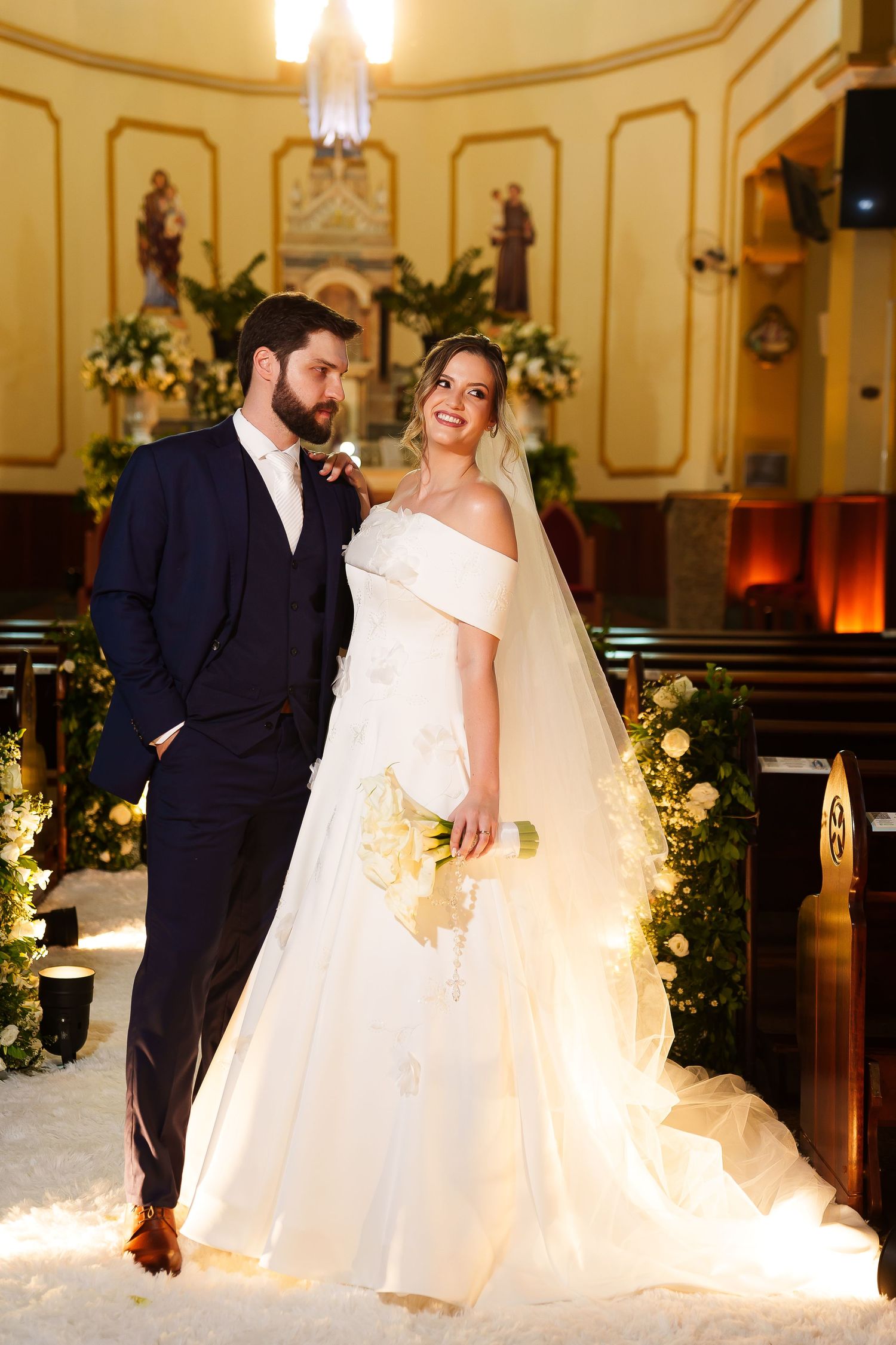 Bride and groom posing together in a decorated church, bride in white off-shoulder gown holding bouquet.