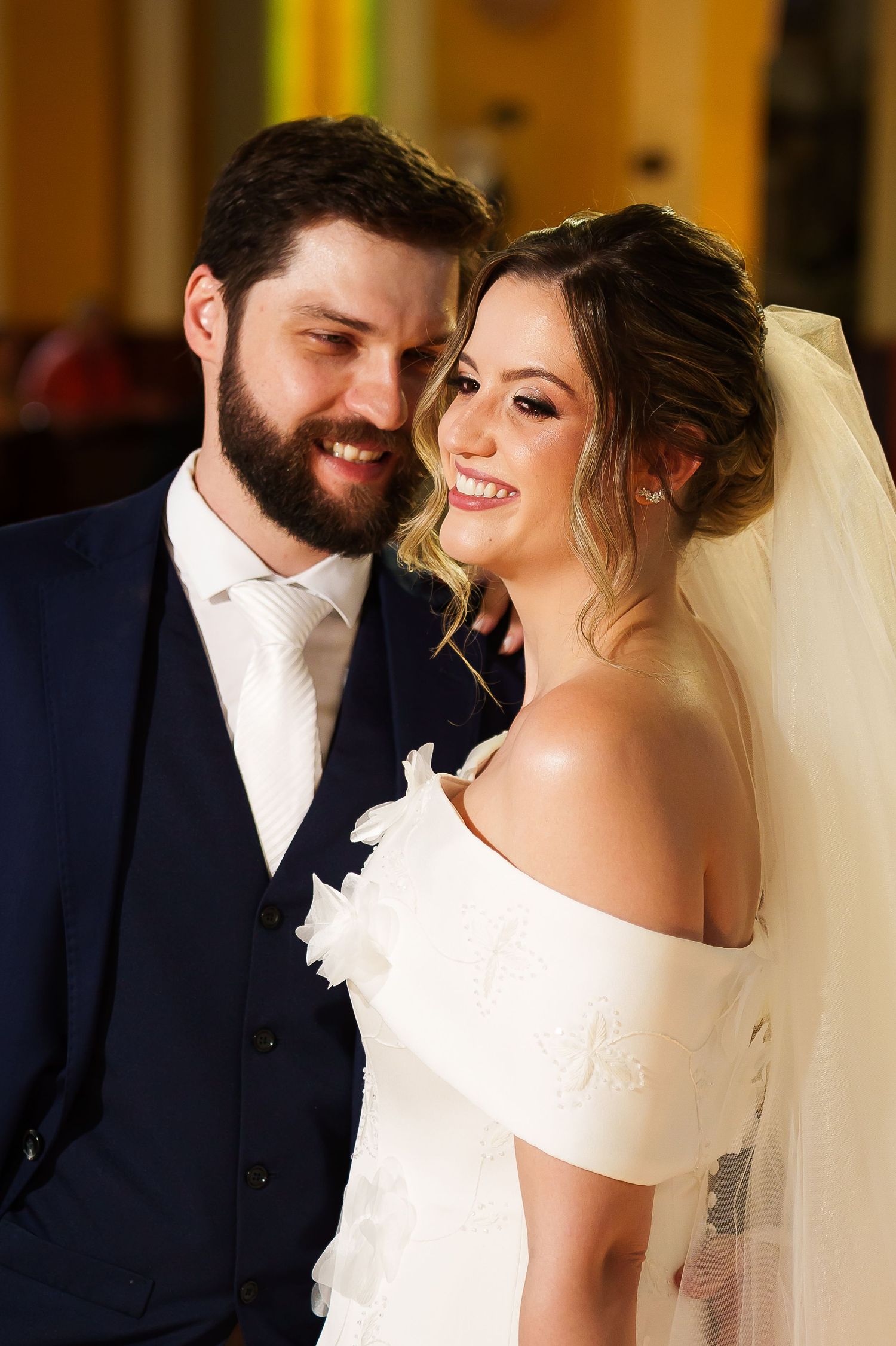 Smiling bride in white off-shoulder gown with veil poses with bearded groom in navy suit at wedding ceremony.