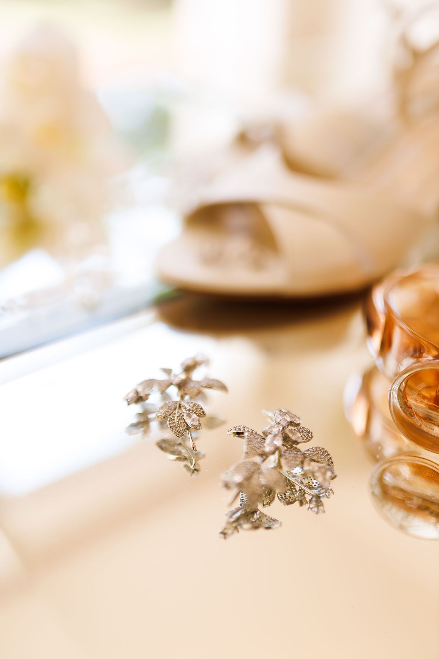 Silver floral bridal earrings on a white surface with heels and perfume bottle in soft bokeh background.