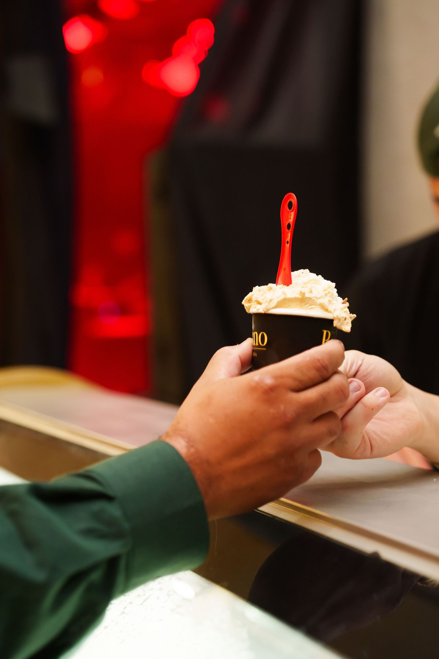 Person in green sleeve receiving a dark cup of ice cream topped with whipped cream and a red spoon at a counter.