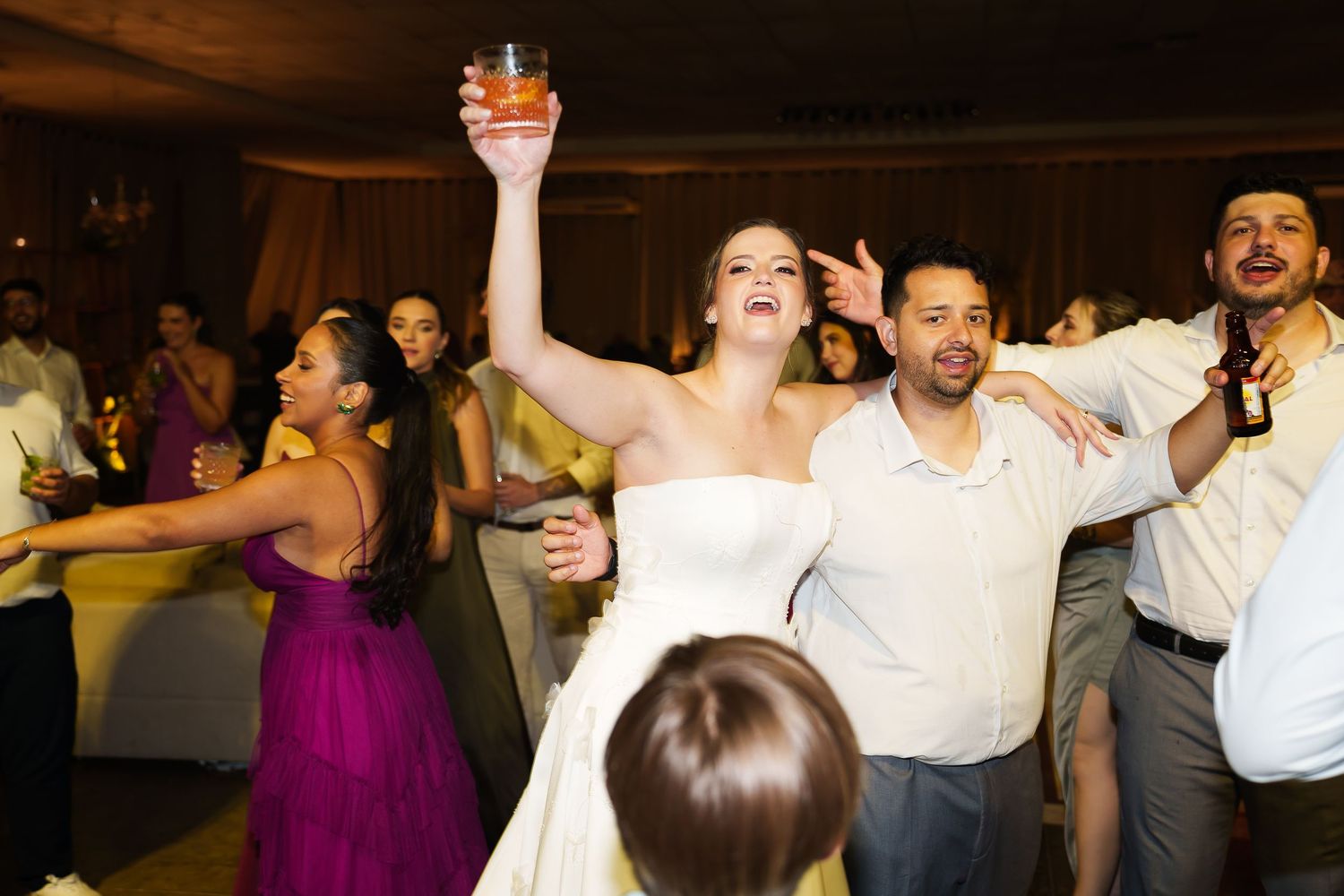 Bride in white gown raises cocktail glass while dancing with guests at elegant wedding reception.