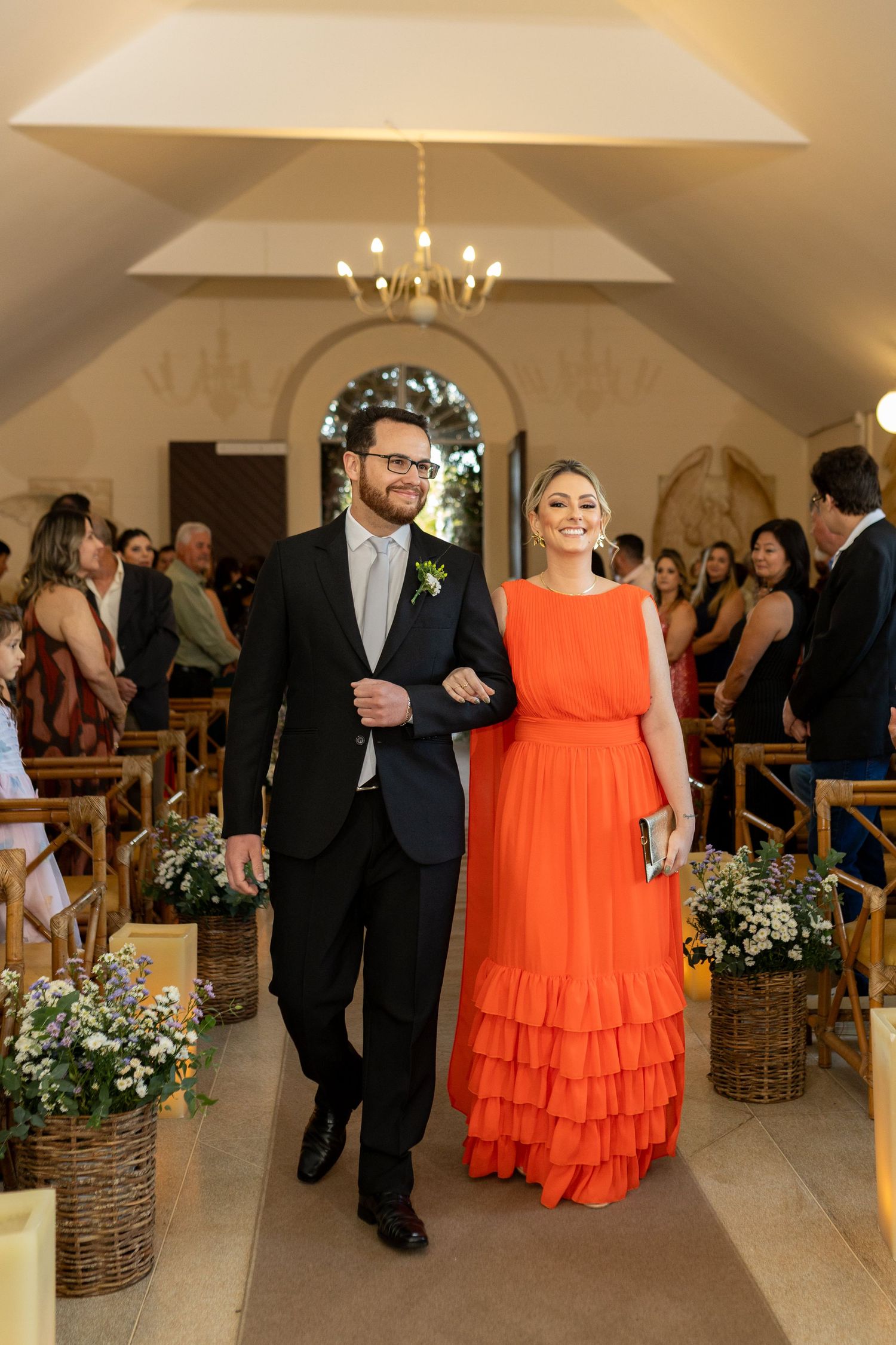 A man in a dark suit and woman in an orange ruffled gown walk down a church aisle at a wedding ceremony.