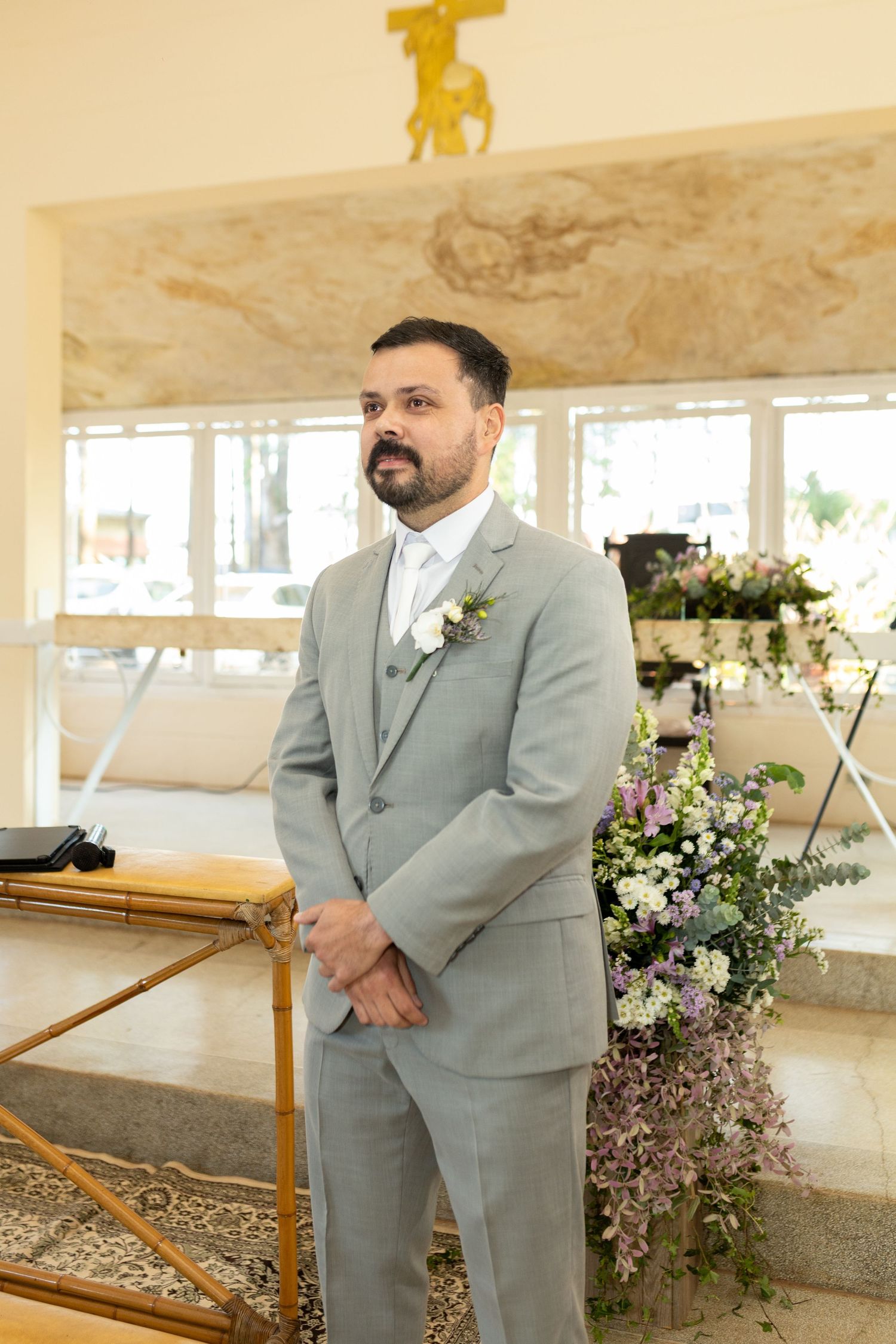 Groom in gray suit with boutonniere stands at wedding altar beside floral arrangement.