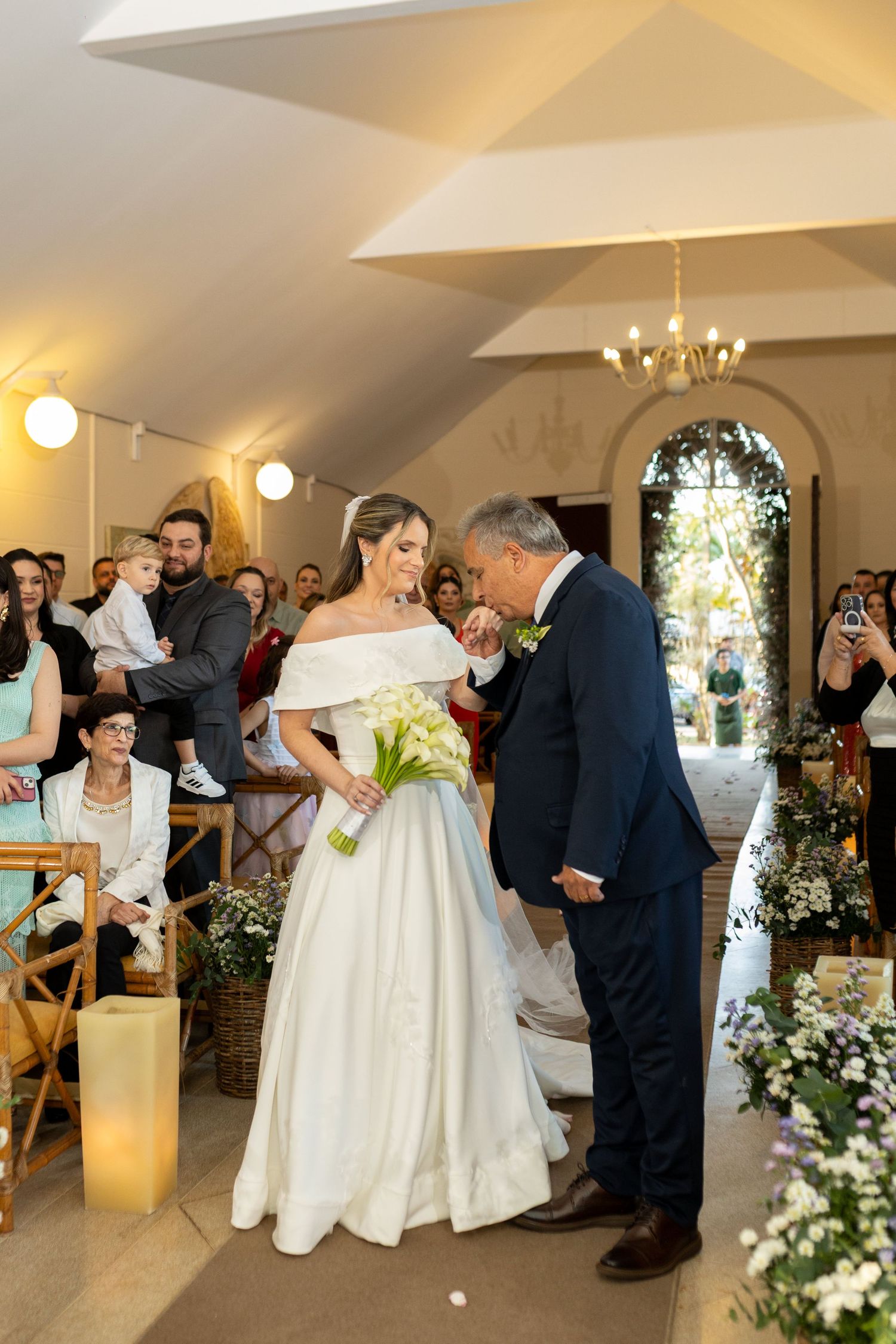 A groom kisses the bride's hand at a wedding ceremony as guests watch in a warmly lit chapel.
