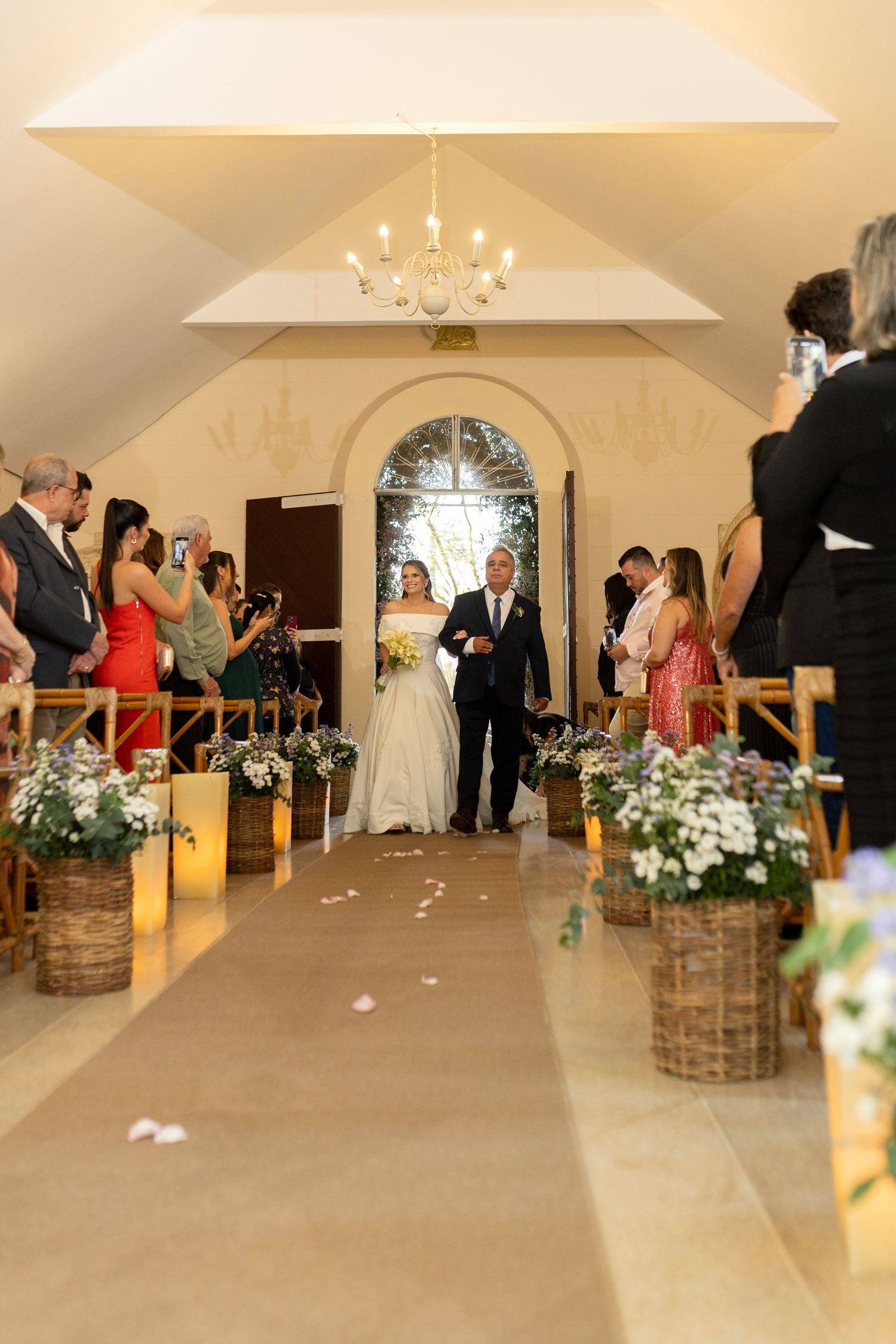 Bride walks down the aisle with escort in elegant chapel, flanked by floral baskets and candlelit decor.