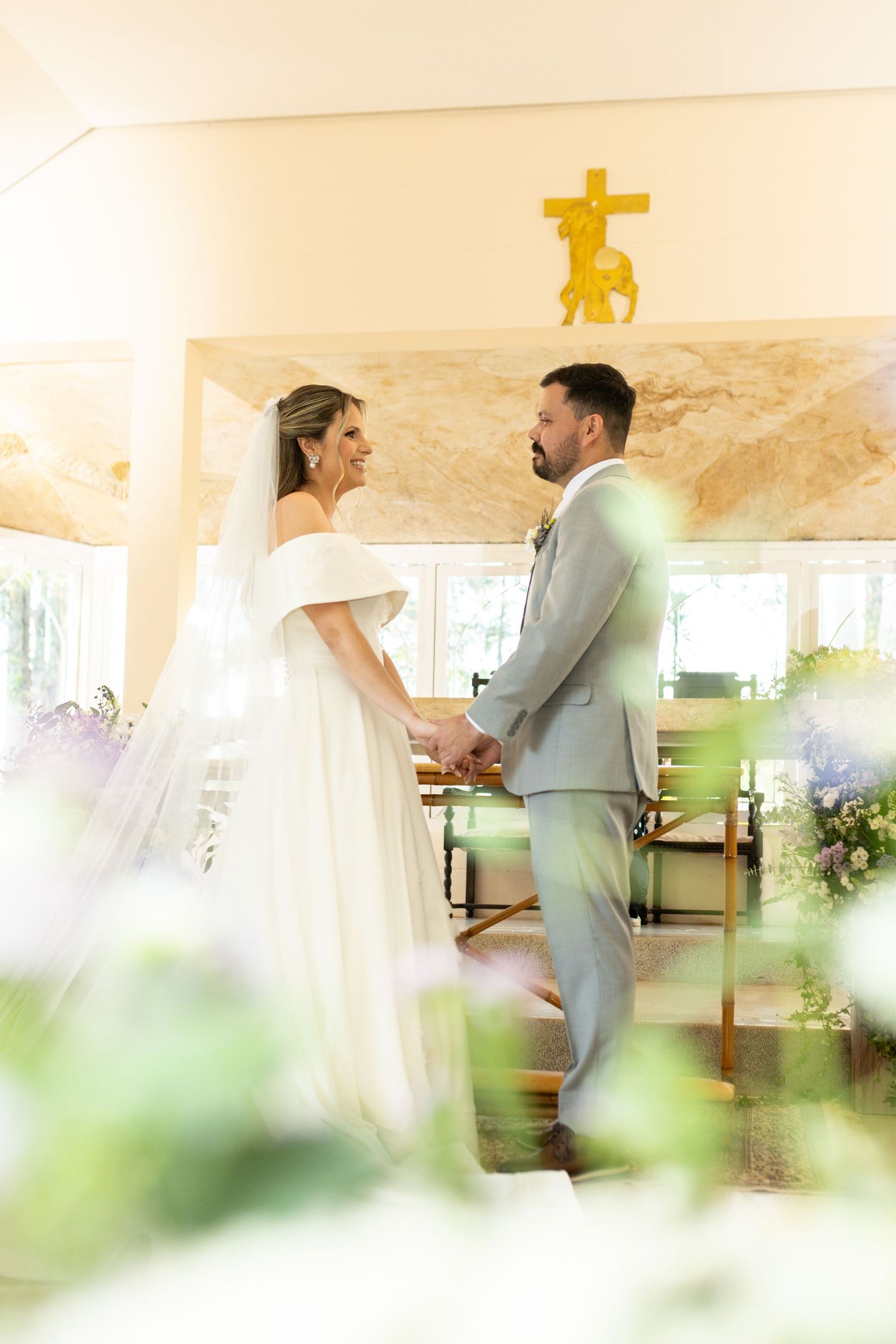 Bride and groom hold hands at altar in sunlit chapel with gold cross, she in white gown, he in gray suit.