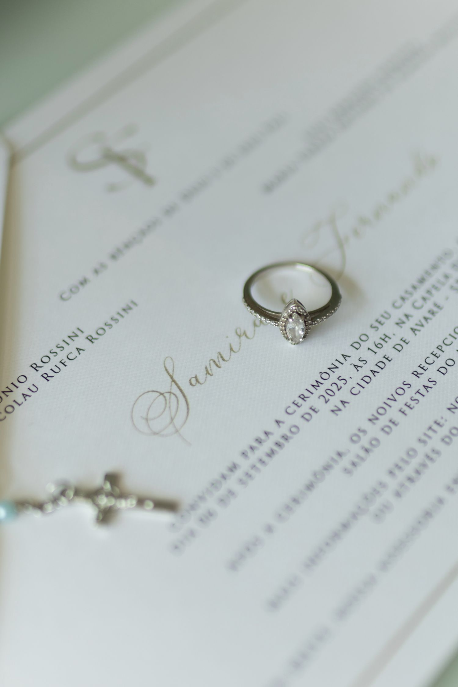 Elegant diamond engagement ring resting on a wedding invitation with gold script, alongside a delicate cross bracelet.
