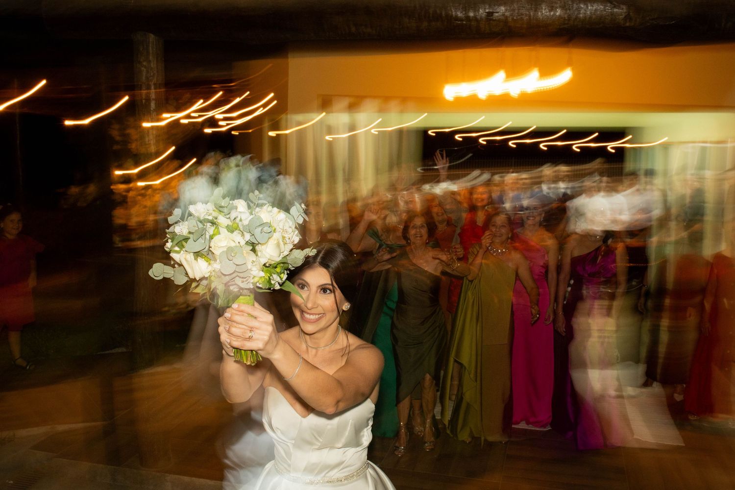 Bride in white gown raises bouquet of white and blue flowers at wedding reception with guests in background.