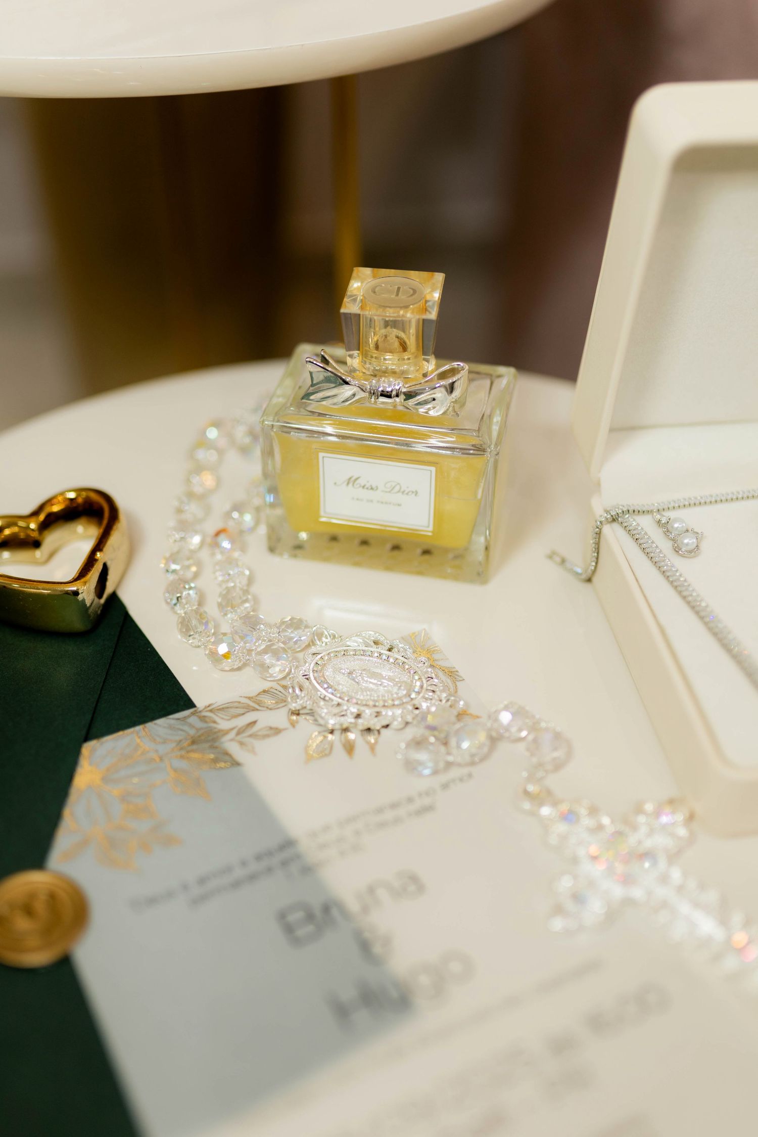 Elegant bridal flat lay featuring Miss Dior perfume, crystal jewelry, gold heart trinket, and wedding invitation on white table.