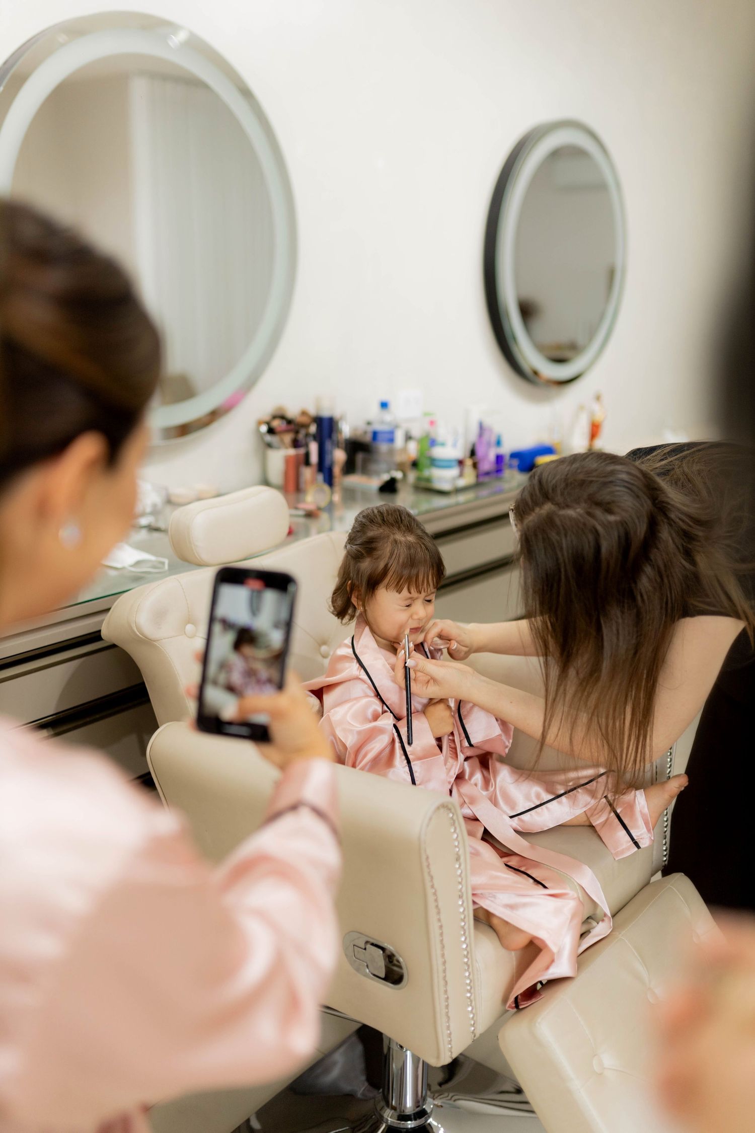 A young girl in a pink robe gets her hair done at a salon while a woman photographs her on a smartphone.