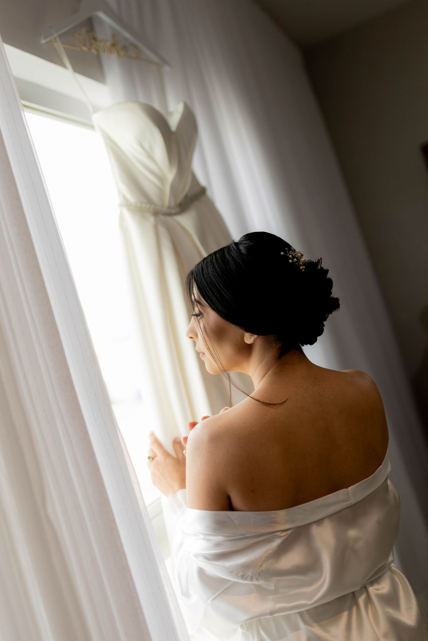 Bride in white robe gazes at elegant wedding dress hanging by window, hair styled in updo with floral accessories.