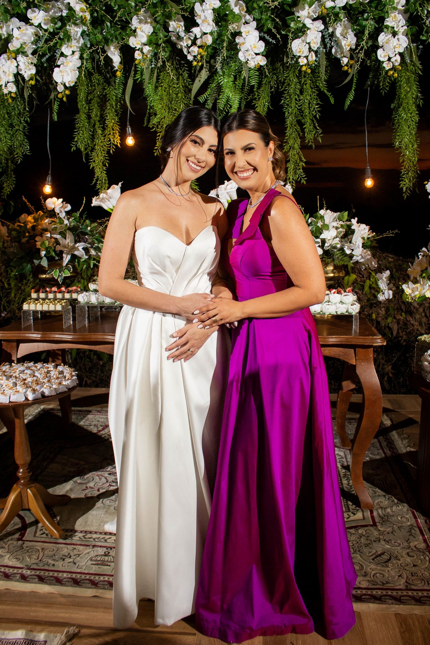 Bride in white gown poses with guest in magenta dress at elegant floral wedding reception.
