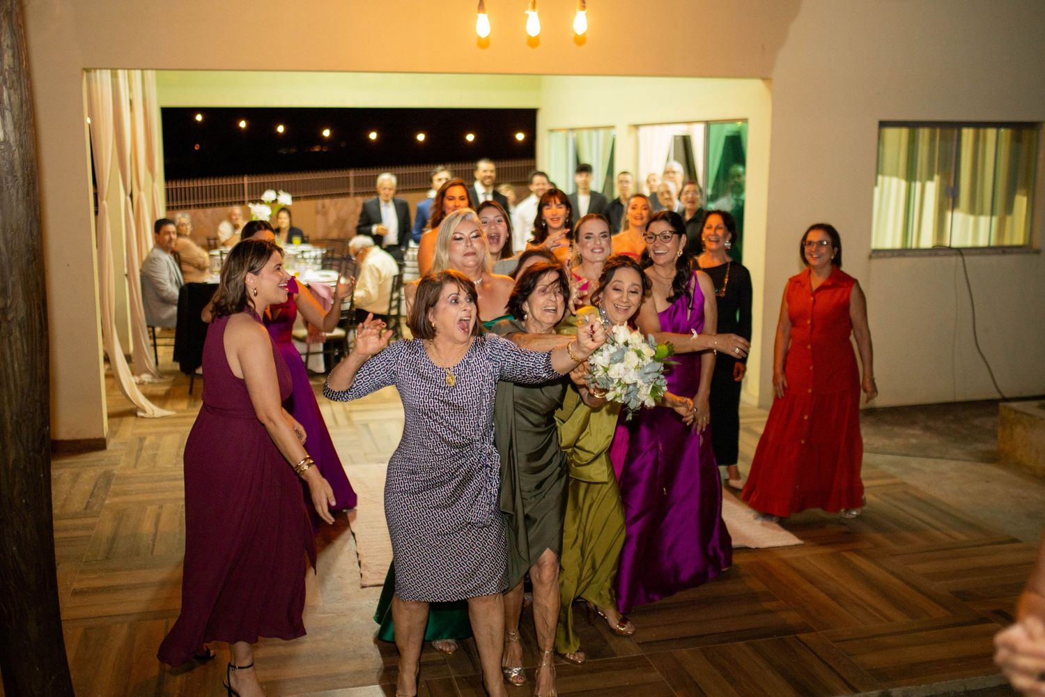Women in colorful dresses eagerly reach for a bridal bouquet toss at an elegant wedding reception.