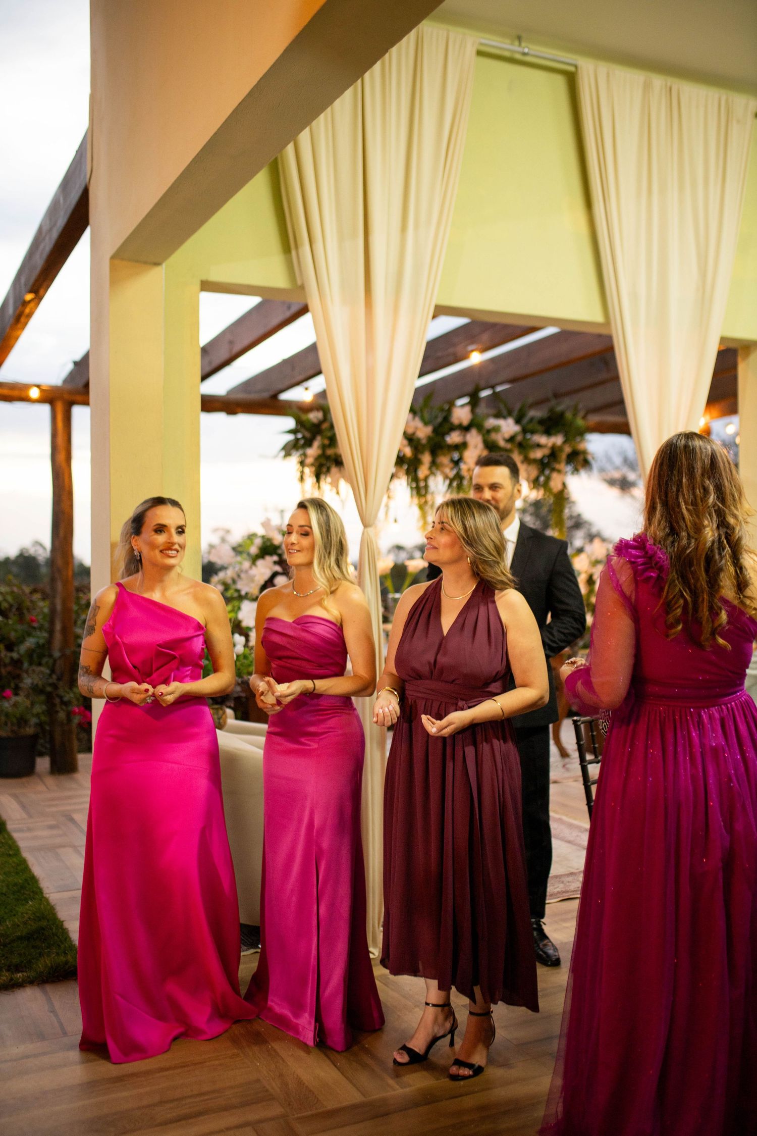 Women in pink and burgundy formal gowns socializing at an elegant outdoor wedding reception under a draped pergola.