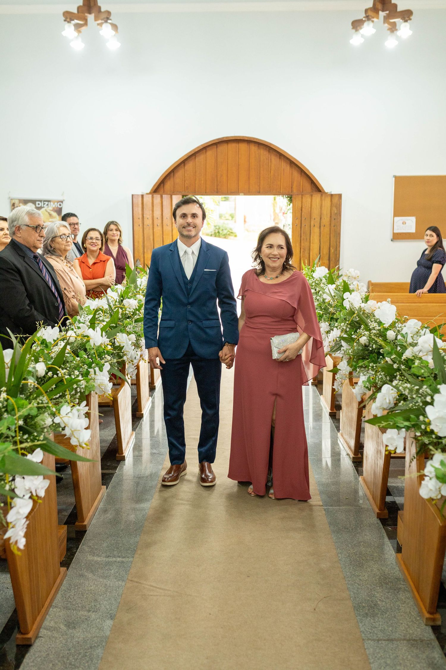 Groom in blue suit walks church aisle with woman in rose dress, flanked by white floral arrangements on wooden pews.